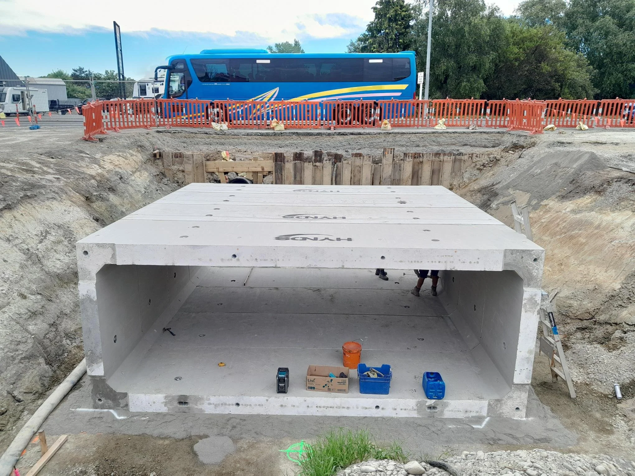 Under construction of a large underground concrete storm drain or utility box with tools inside, at a construction site with excavation and safety barriers in the background.