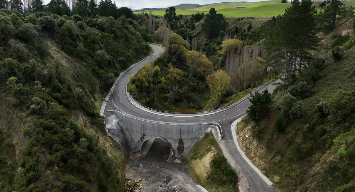 A winding mountain road with a bridge over a small river or creek, surrounded by lush green trees and hills.