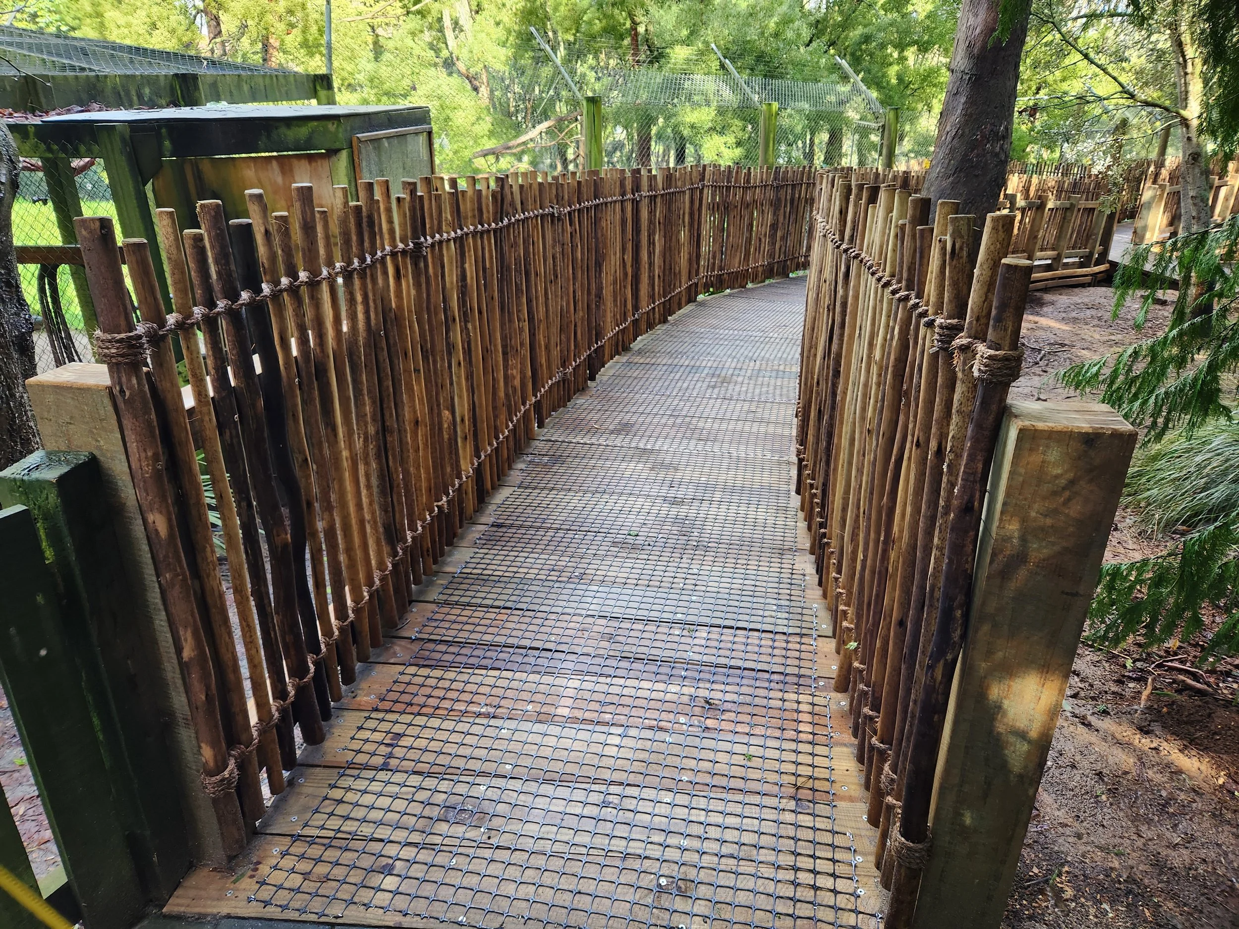 Wooden and wire mesh footbridge in a park surrounded by trees and fencing.