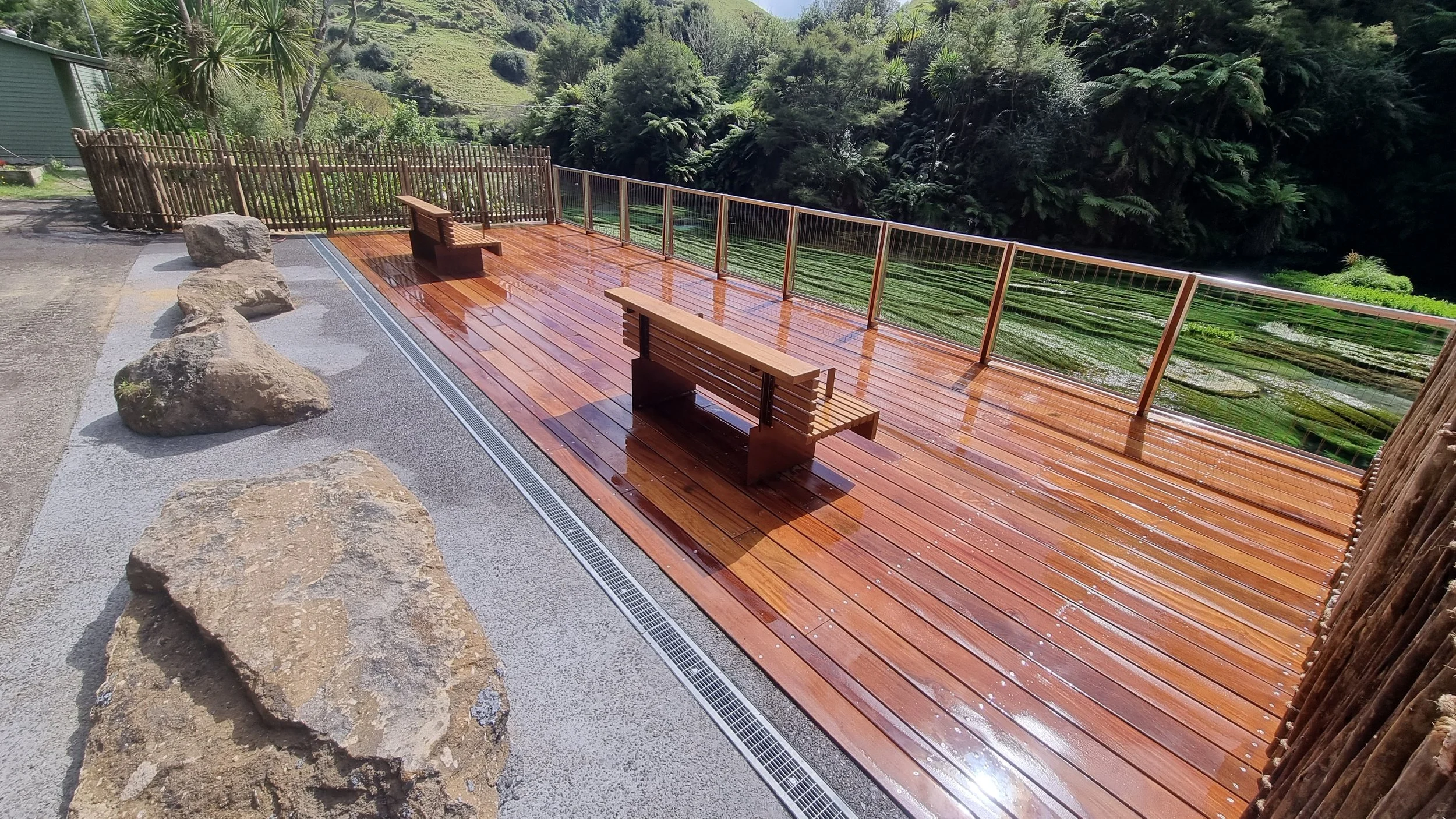 A wooden deck with two benches, surrounded by rocks and a bamboo fence, overlooking lush green trees and hills.