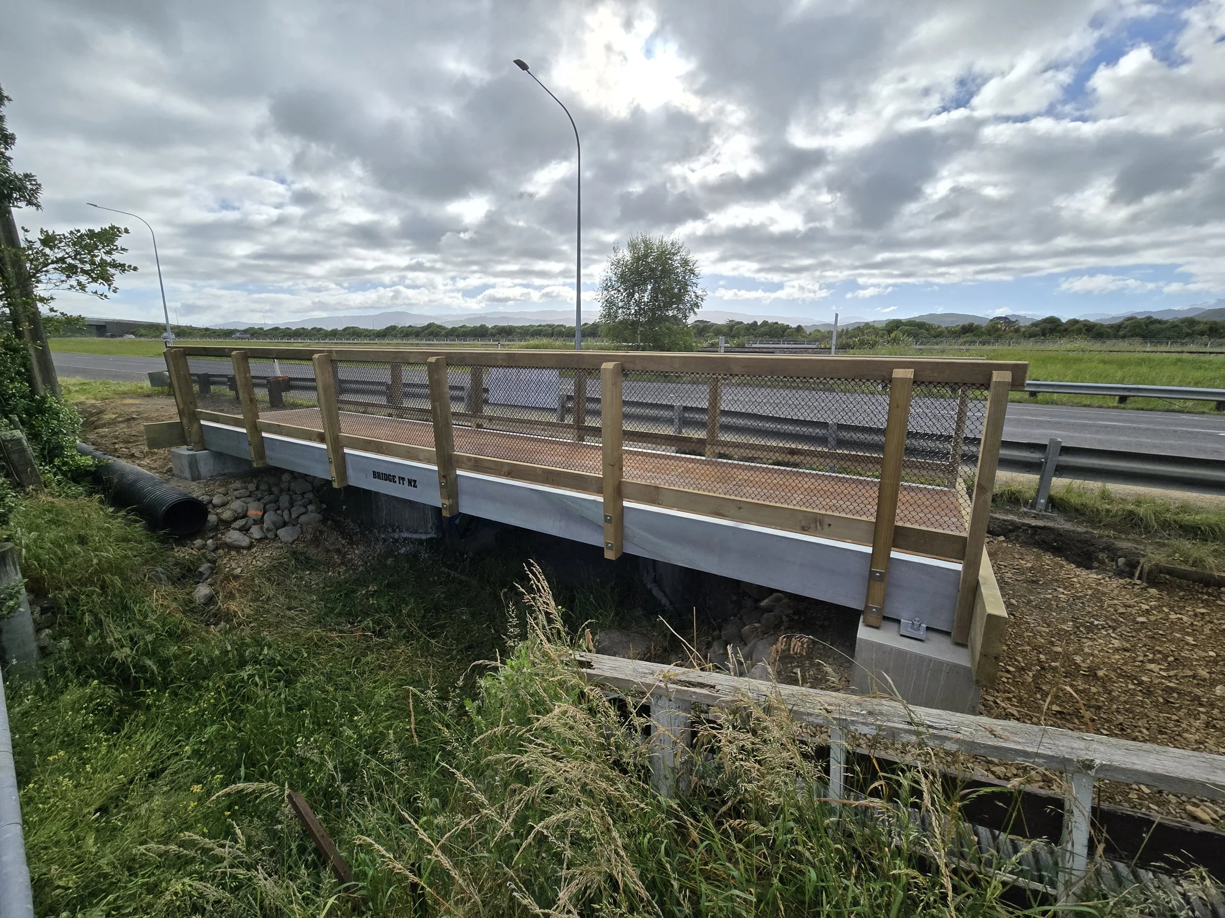 A small bridge with wooden railings over a creek next to a highway with street lamps, trees, and distant hills under a partly cloudy sky.