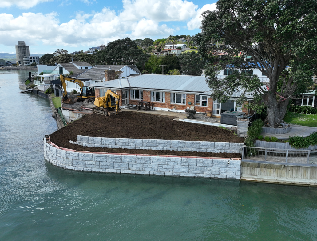 Under construction yard on waterfront with excavators, soil, retaining wall, and house nearby.
