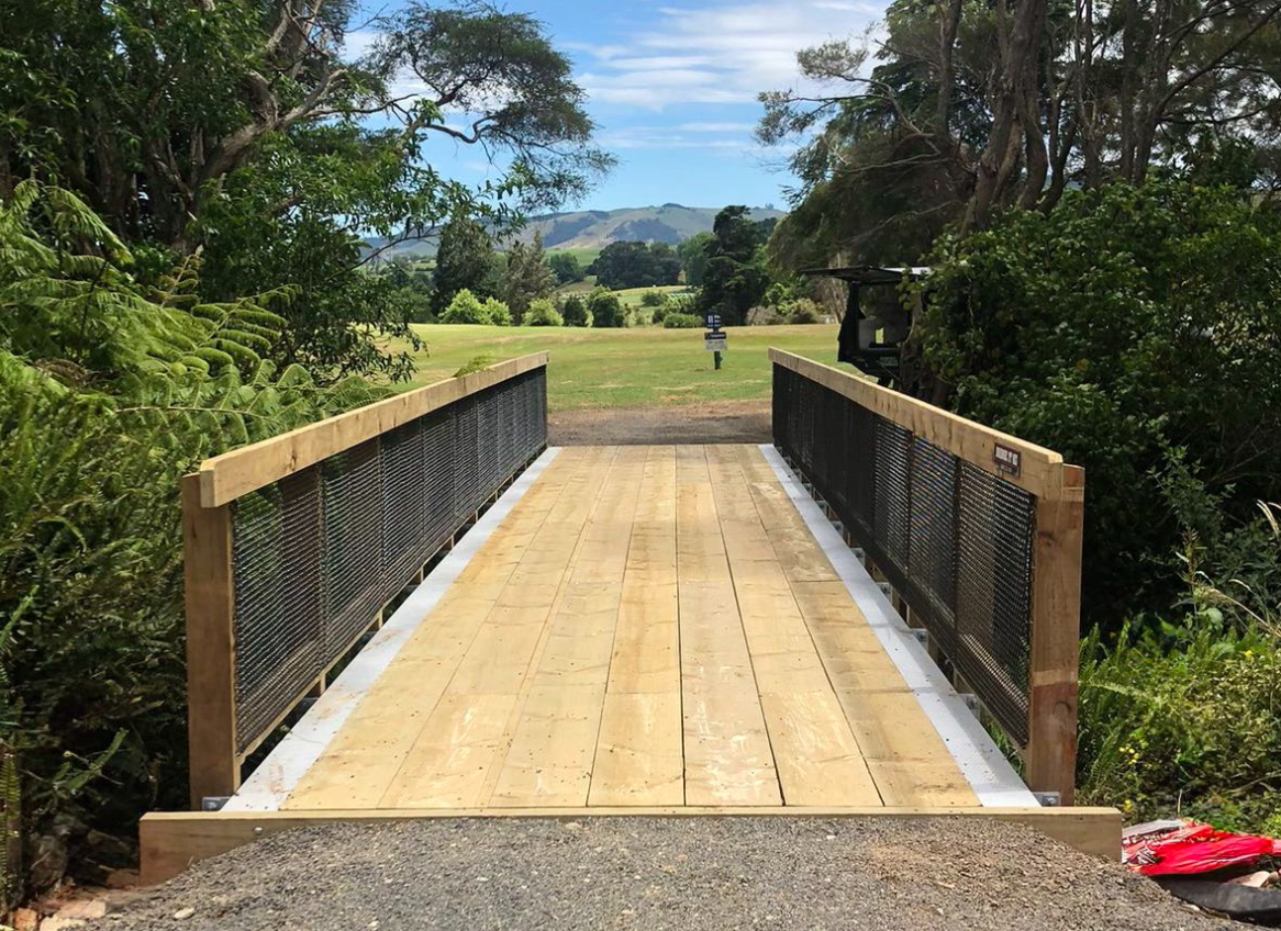 A wooden pedestrian bridge with metal railings over a small creek, leading to a grassy area with trees and rolling hills in the background under a partly cloudy sky.