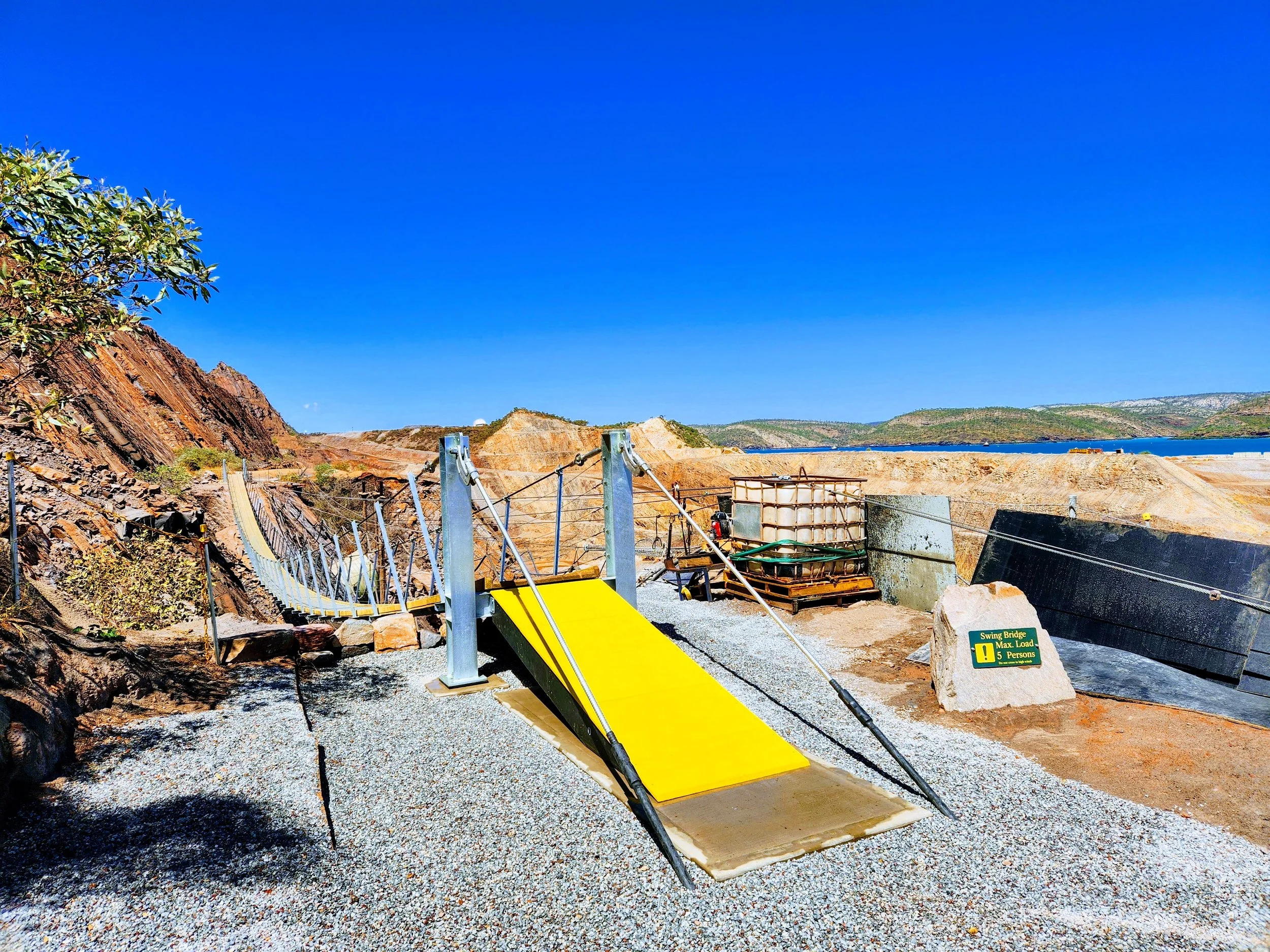 A suspension bridge with a yellow walking surface under a clear blue sky in a rocky outdoor area with hills.