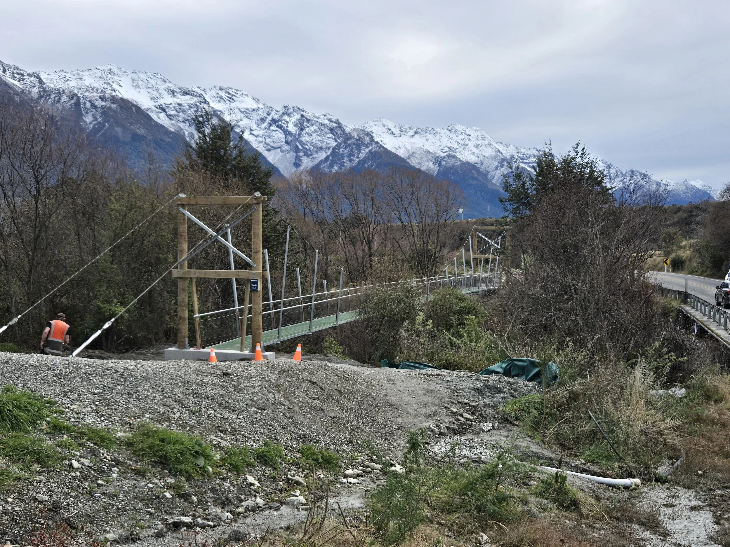 A partially constructed pedestrian suspension bridge over a rocky and muddy terrain with snow-capped mountains in the background.