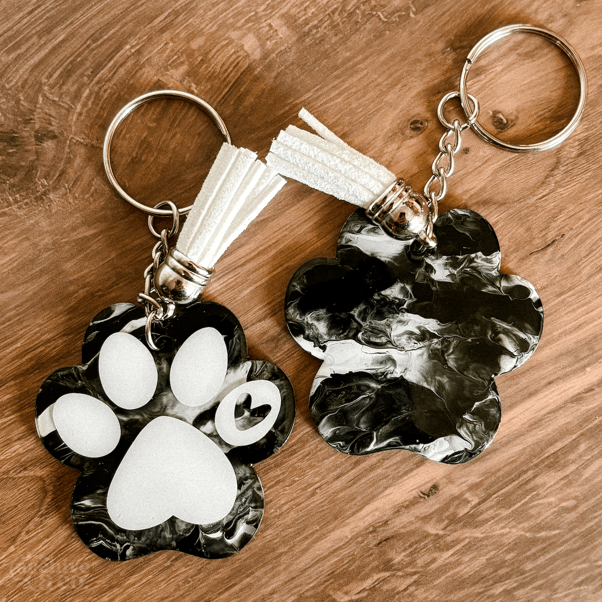two handmade paw print keychains with black and white marbled patterns, white tassels, silver keyrings, and resin finish on a wood background