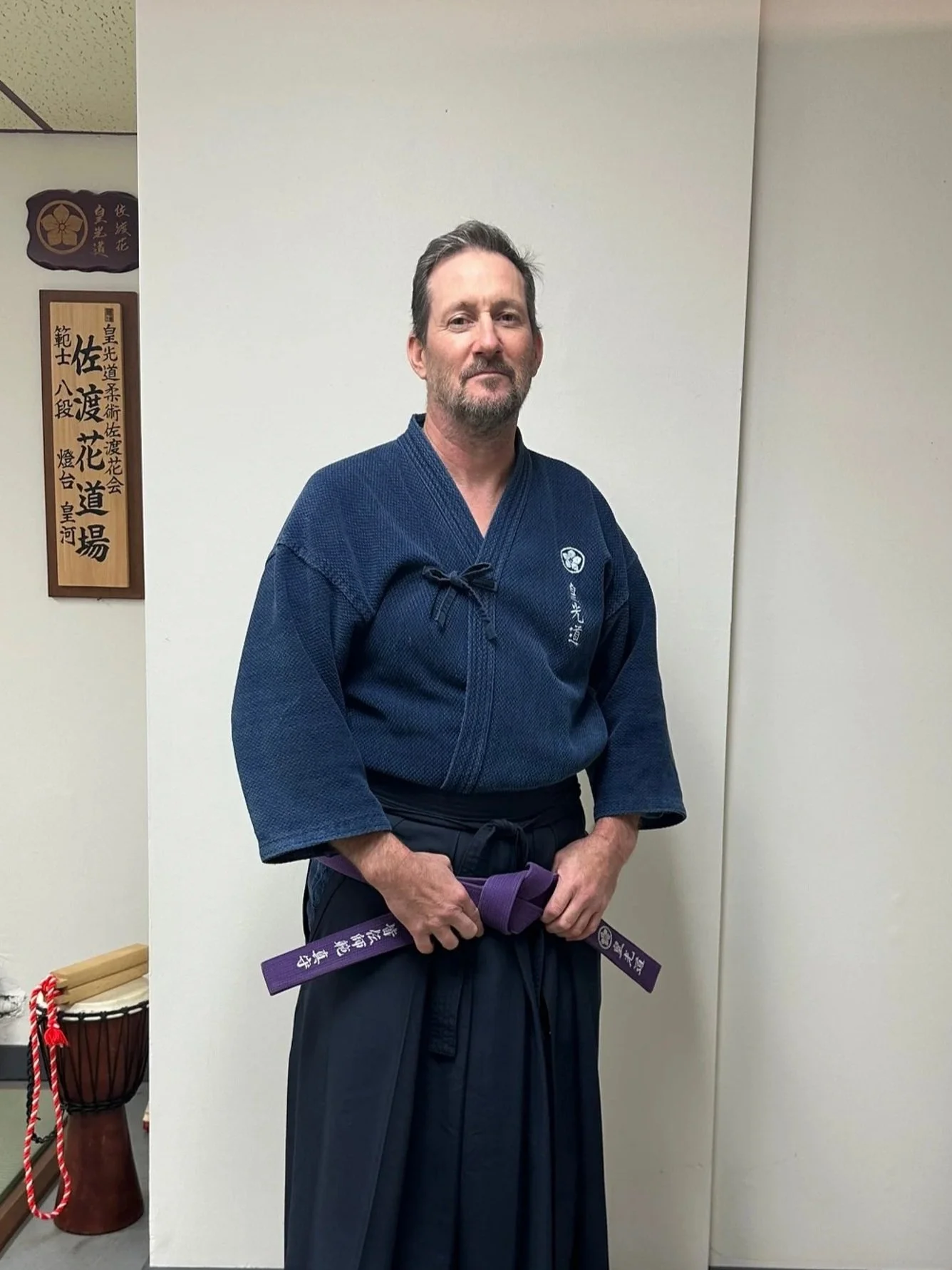 Two martial artists practicing KoKoDo JuJutsu on a dojo mat, one lifting the other in a white gi with black belt, in a training hall with wooden walls and a decorative framed artwork in the background.