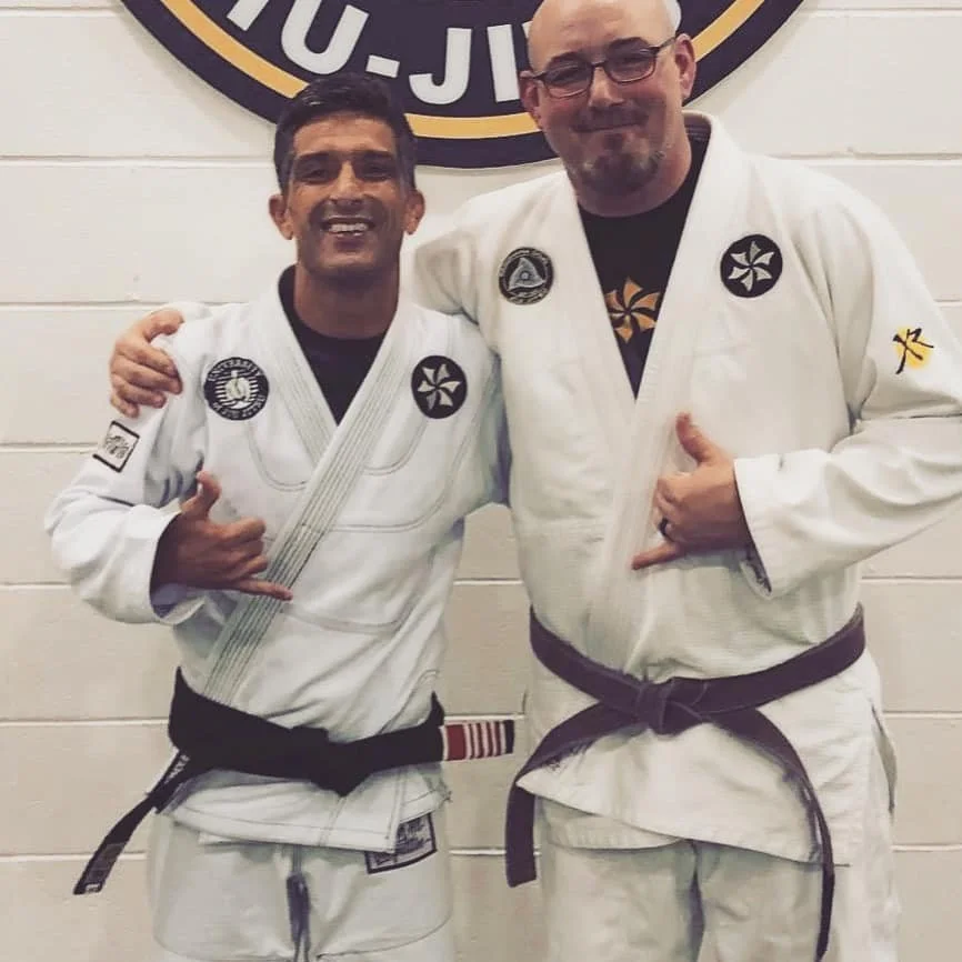 Pagani Sensei and Michael Seamark white Brazilian Jiu-Jitsu Six blades gis posing together in front of a wall with a circular logo. They are smiling and making hand gestures, with one giving a thumbs-up and the other inside a fist.