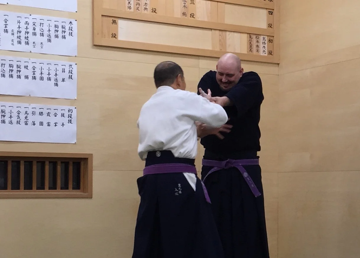 Two martial artists practicing kokodo jujutsu in hombu dojo, one wearing a white gi and purple belt, and the other in a black gi with purple belt, engaged in a demonstration of technique.