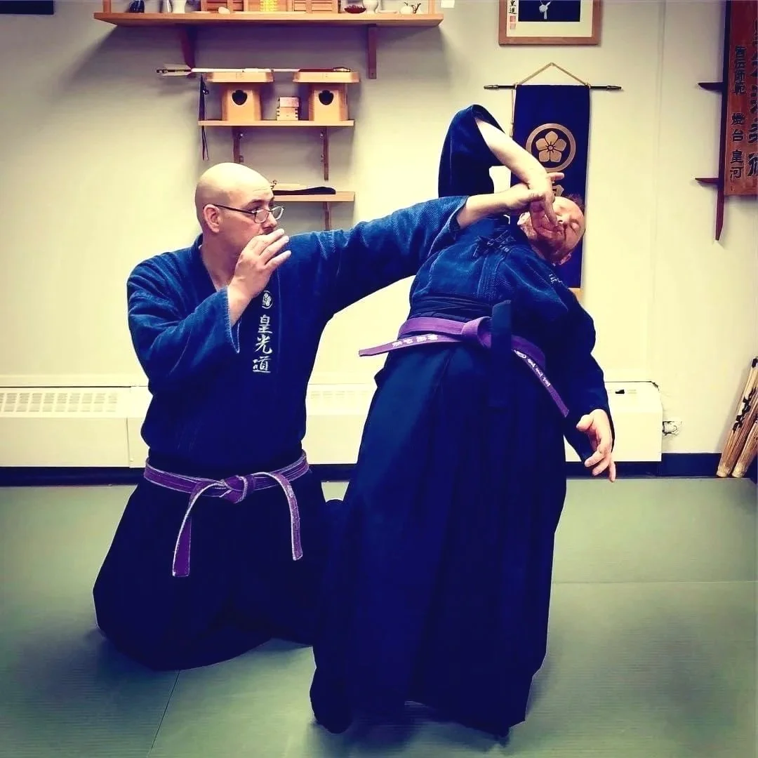 Two martial artists practicing KoKoDo JuJutsu in sadohana dojo vancouver bc. One person is applying a technique to the other, who is leaning forward with their head turned to the side. Both wear traditional various colored gi uniforms.