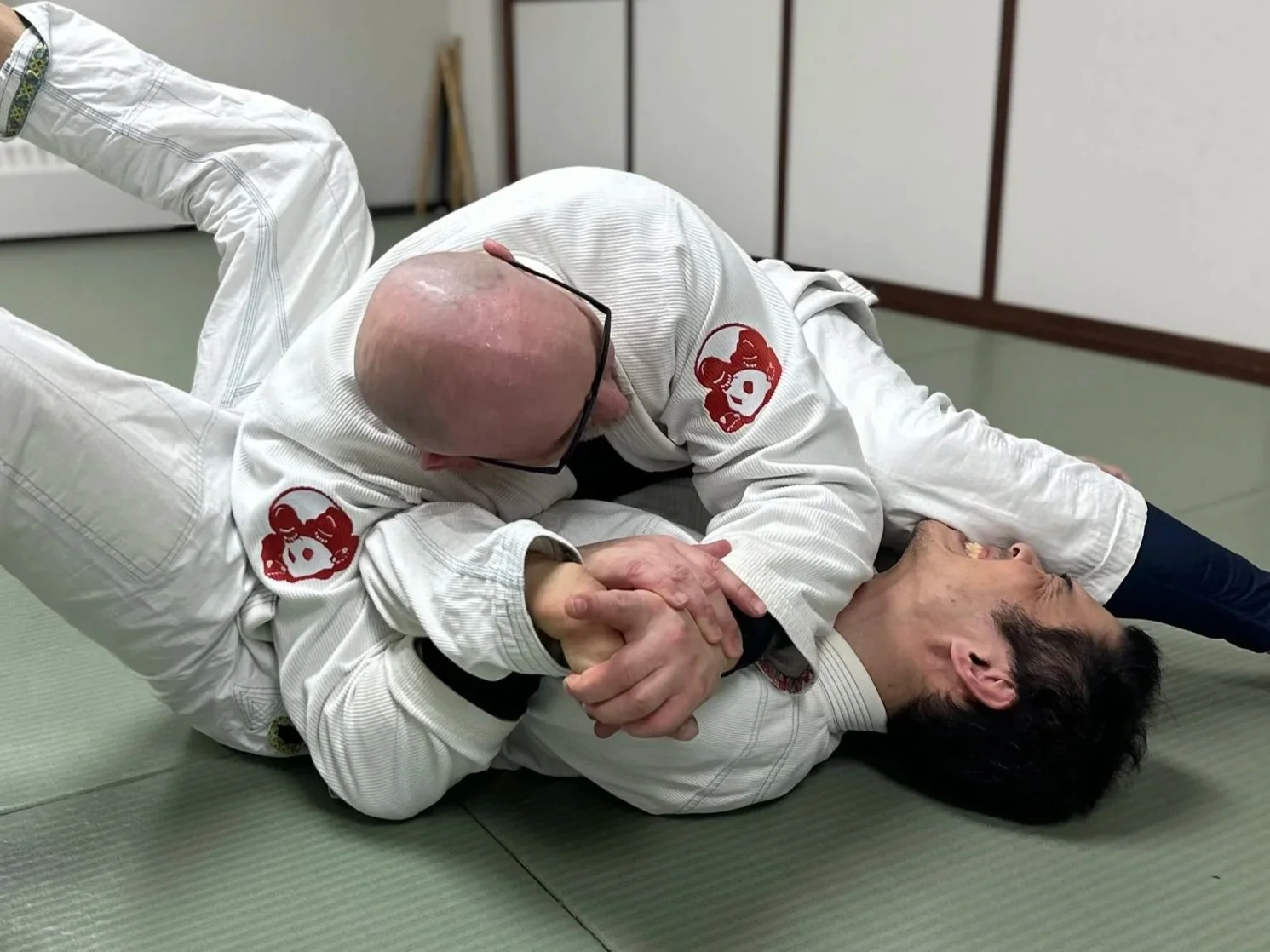 Two jiu-jitsu practitioners in white gis with red logos on the shoulders, engaged in a grappling technique on a mat indoors. One person is applying a chokehold on the other.