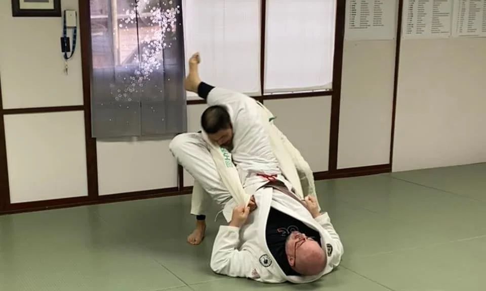 Two martial artists practicing Brazilian Jiu-Jitsu, one on top and the other on the bottom, in a gym with mats and wall decor.