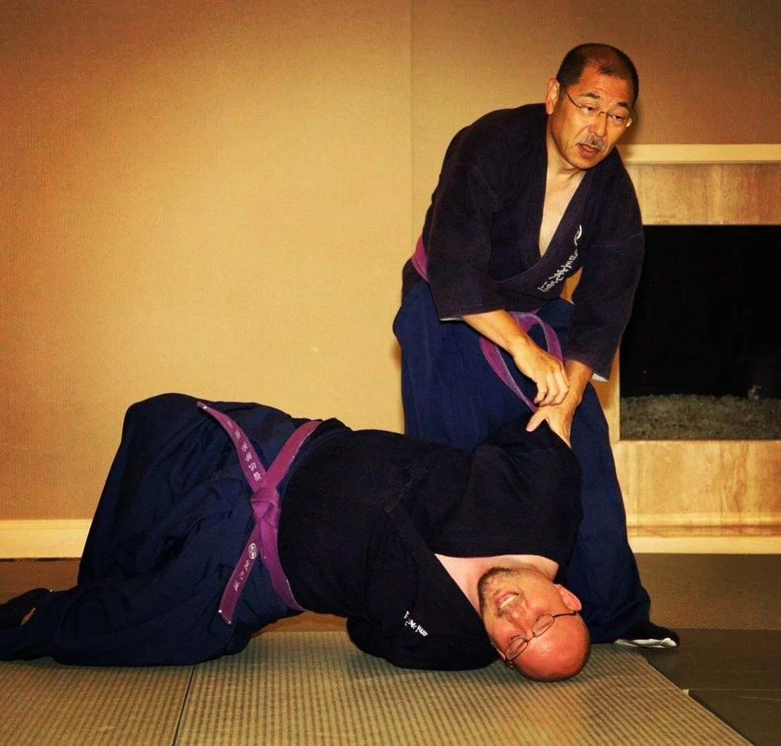 Hanshi and Soke practicing kokodo jujutsu one is in a kneeling position, the other is lying on the mat with glasses, smiling, and being controlled by the Soke Irie Yasuhiro