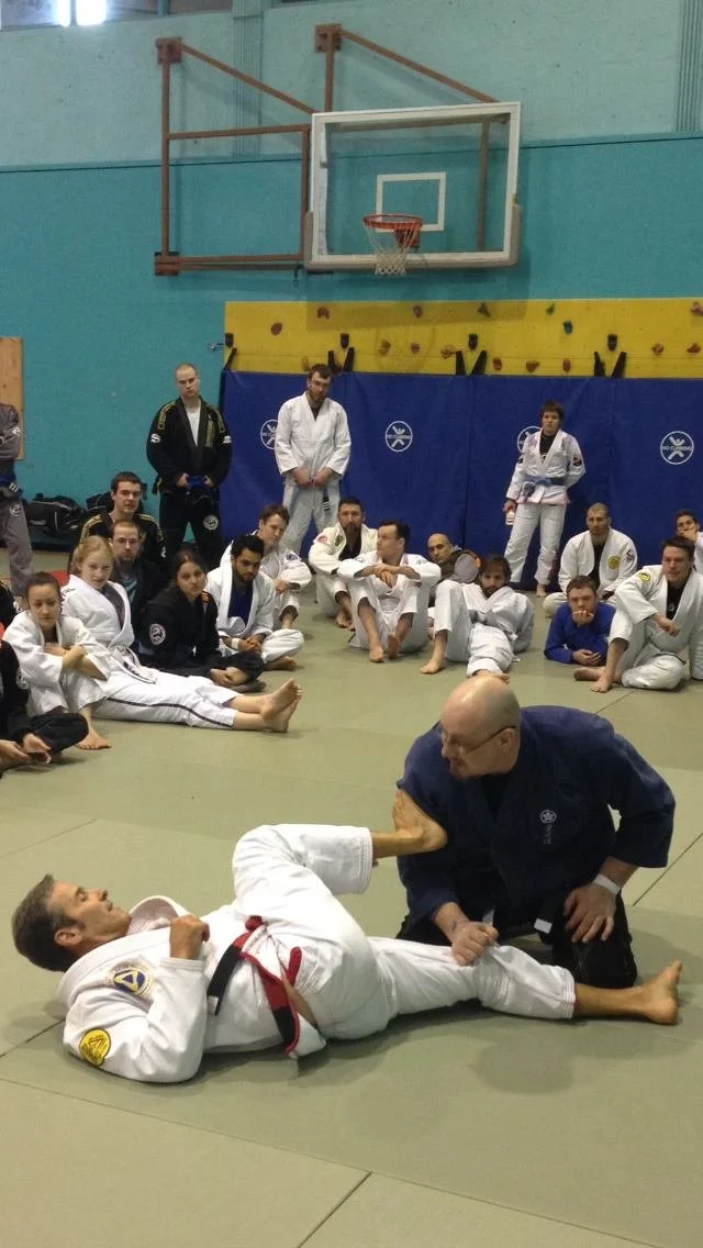 Brazilian Jiu-Jitsu class practicing techniques on the mat with students in gi uniforms, a coach demonstrating a move with a participant, and a group of students observing in a gymnasium with a basketball hoop overhead.