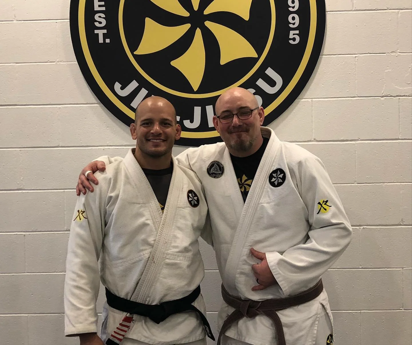 Xande Ribeiro and michael seamark in Brazilian jiu-jitsu uniforms standing in front of a wall with a large martial arts curucuruland bjj emblem. They are smiling, with one man having his arm around the other.