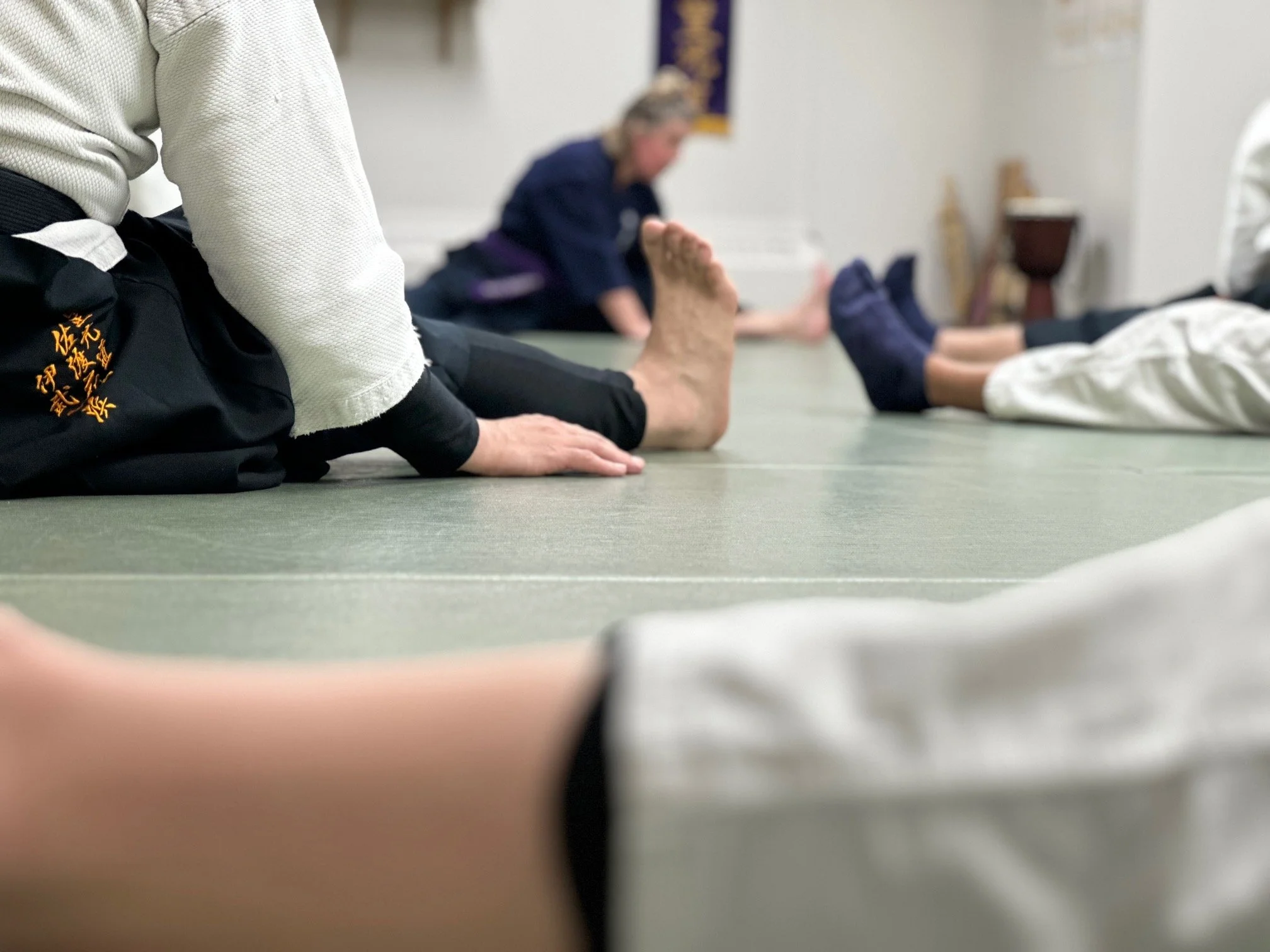 Shihan Diane Seamark is showing students yoga for jiujitsu in vancouver while practicing stretching exercises on a gray mat, seated on their knees or lying on their sides in a dojo or training room.