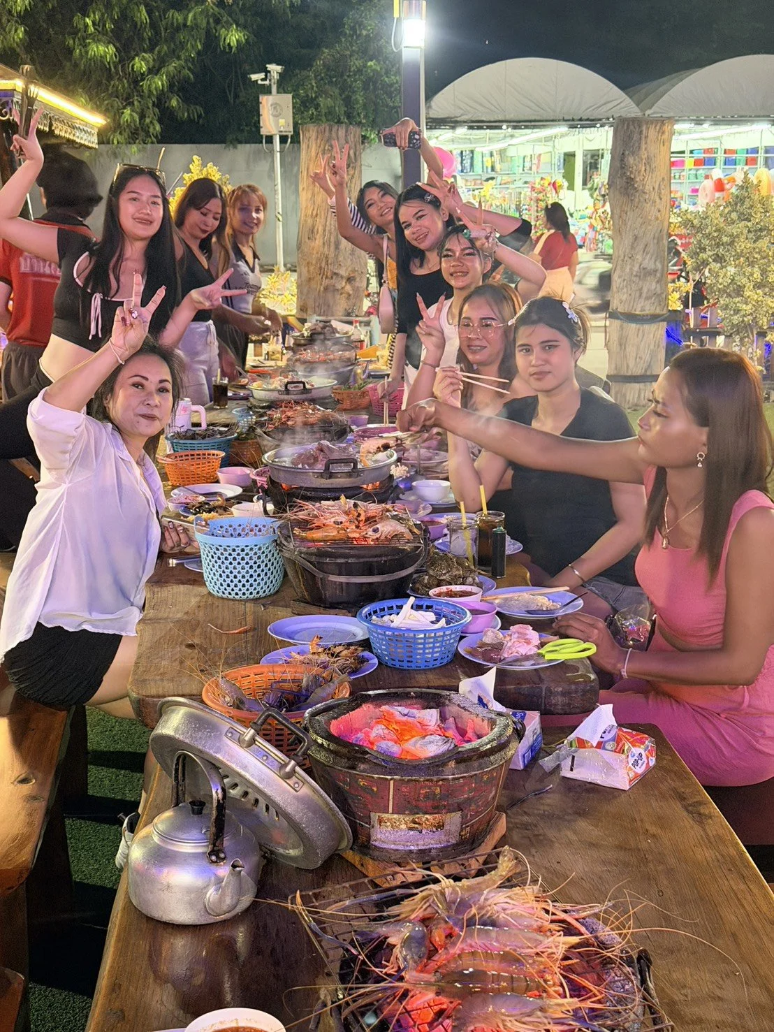 Group of people sitting and standing around a long outdoor table filled with seafood and hot pots, celebrating a gathering at night, with colorful lights and a food stand in the background.
