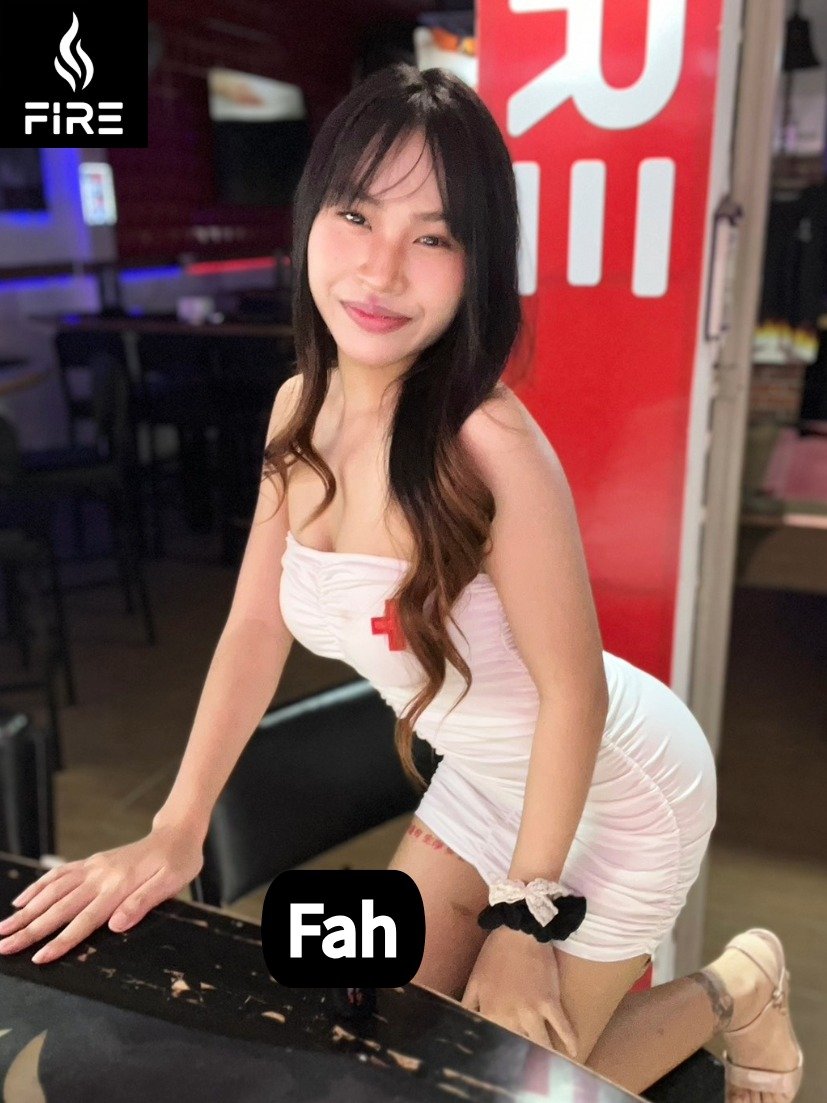 A woman in a white nurse costume with a red cross logo, posing with one knee and hand on a table in a dimly lit setting, with a fire station sign in the background.