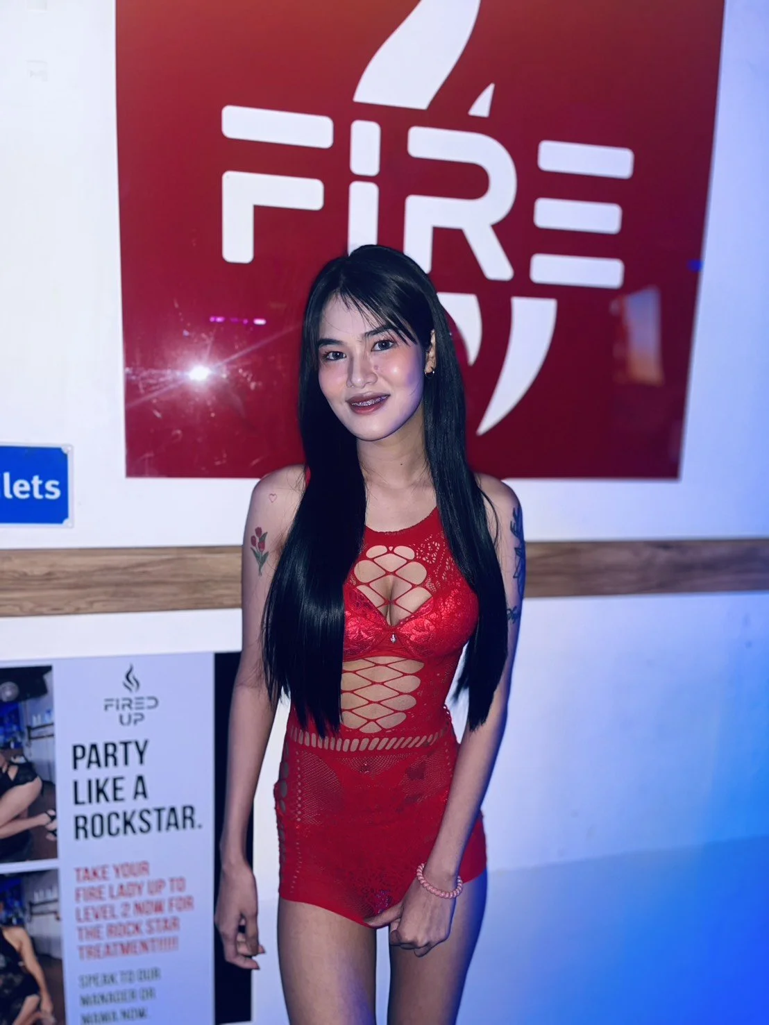 A young woman with long black hair and tattoos on her arms, wearing a red lace dress, standing in front of a fire safety sign at a party.