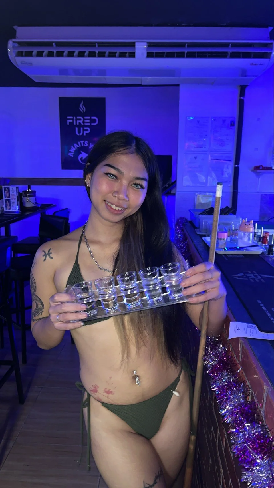 A young woman in a bikini holding a tasting tray of shot glasses and an ouija board stick inside a bar with blue lighting.