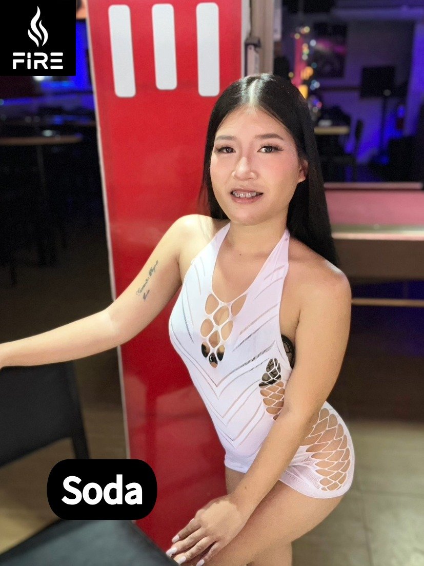 A young woman with long black hair, wearing a revealing white cutout dress, is posing in a dimly lit nightclub or lounge.