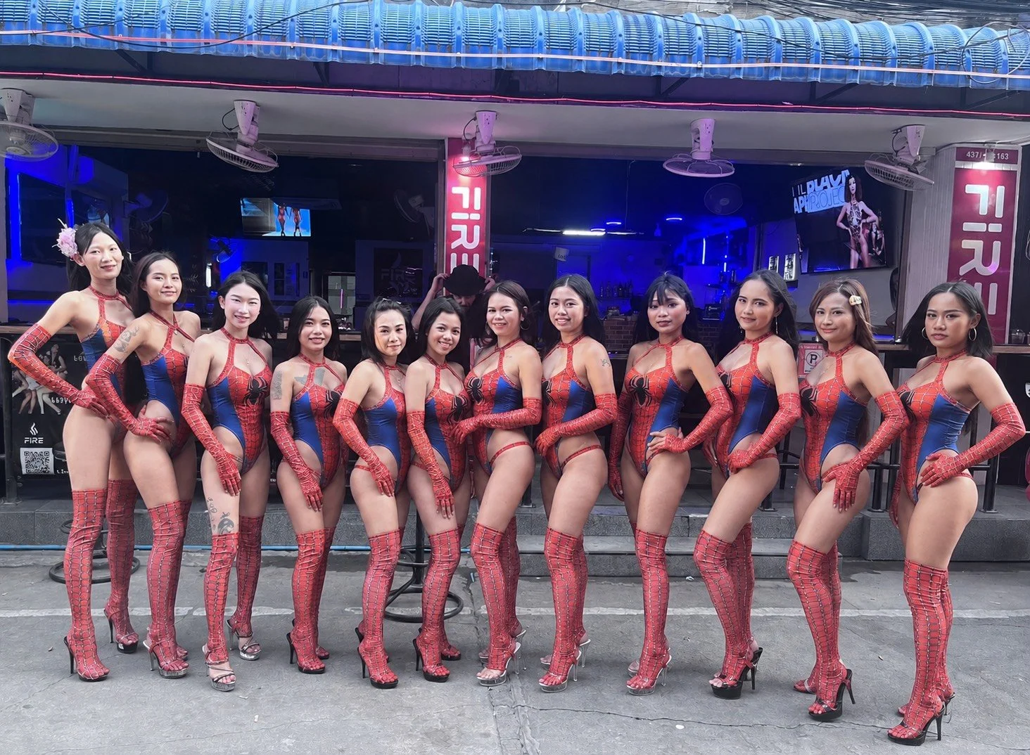A group of 13 women dressed in matching Spider-Man themed costumes, standing together in a line outside a bar or club at night.