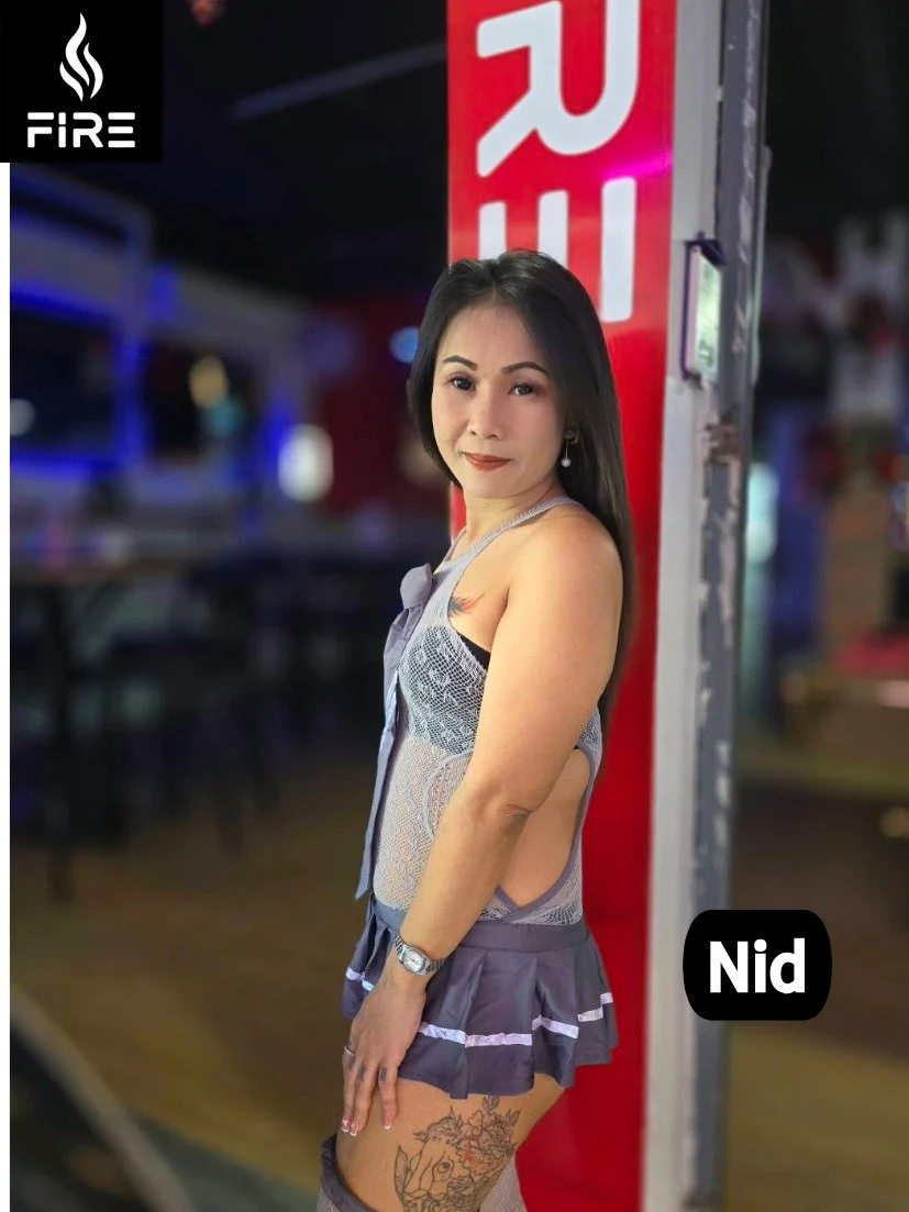 A woman with dark hair posing at a nightclub or bar, standing next to a red pillar with the letter 'R' visible, wearing a revealing outfit with a tattoo on her thigh, and winking at the camera.