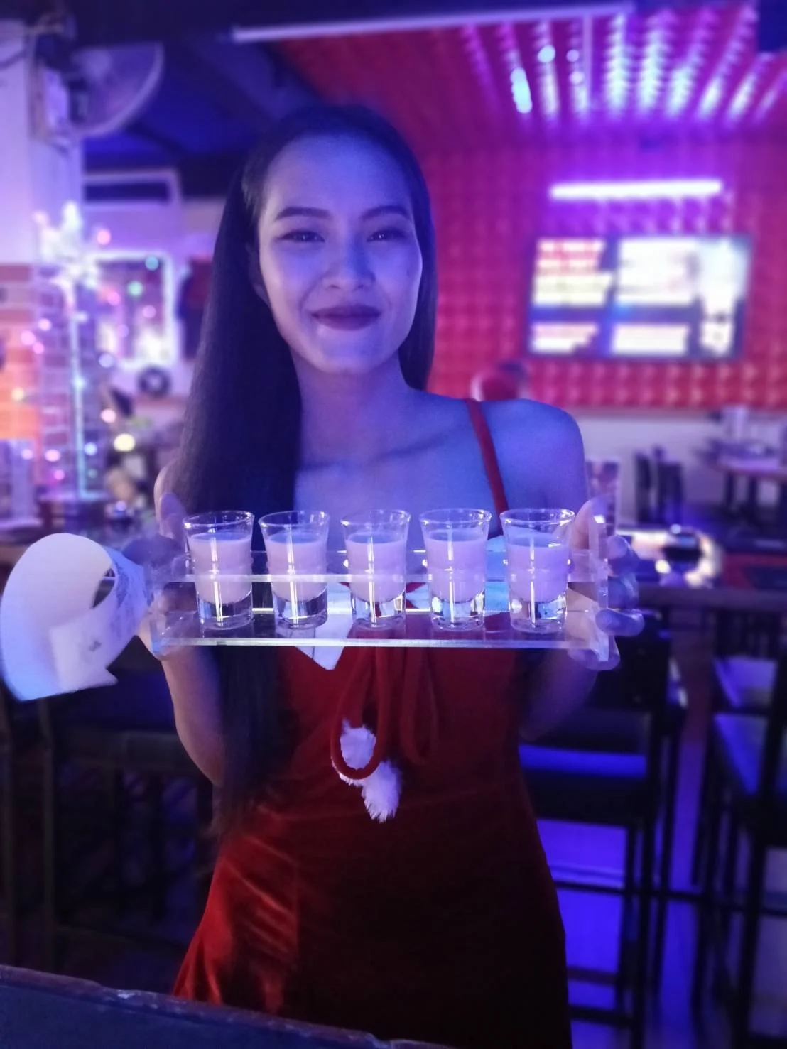 A woman dressed in a red outfit holding a tray with six shot glasses, standing in a dimly lit bar or nightclub with neon purple and red lighting.