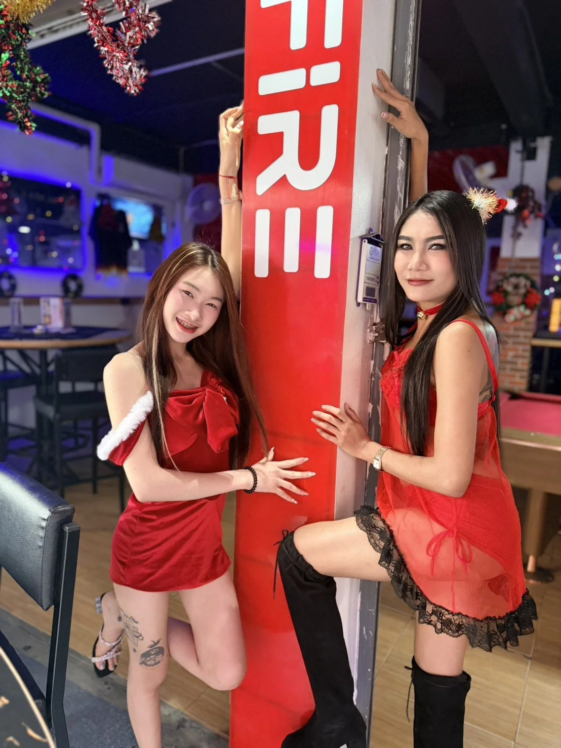 Two women dressed in revealing red outfits posing with a large red and white sign that says 'Hula' inside a festive decorated restaurant or bar.