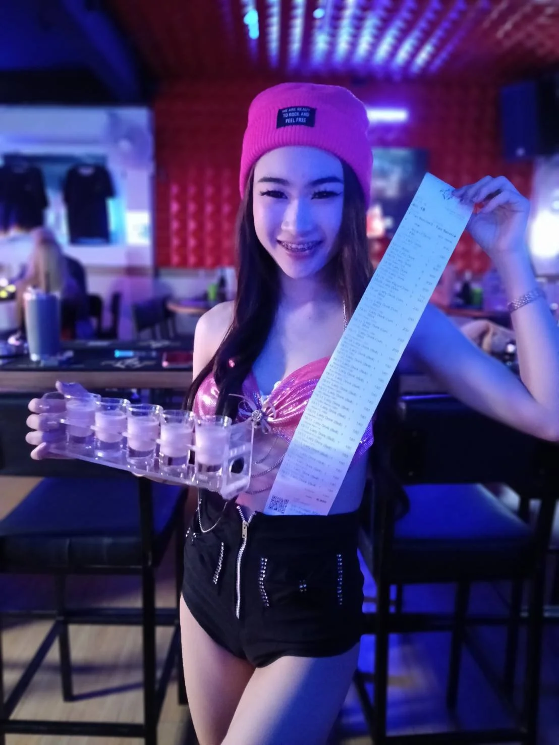 A smiling woman wearing a pink beanie and black shorts holding a long receipt and a tray of shot glasses in a bar with colorful lighting.