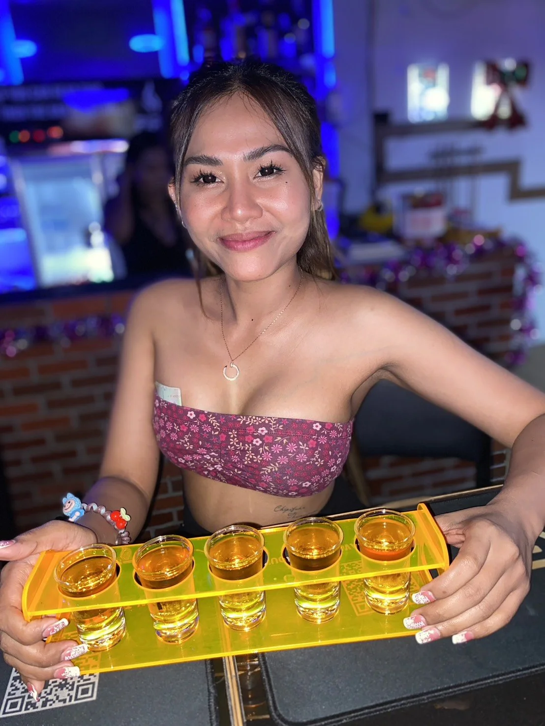 A woman with dark hair smiling and holding a yellow tray of six shot glasses filled with a golden beverage in a bar or restaurant setting.