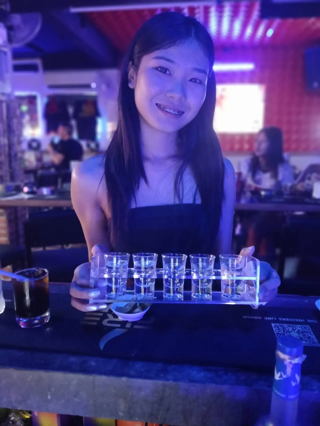 A smiling woman holding a tray with shot glasses filled with clear liquid in a dimly lit bar or club with purple and blue lighting.