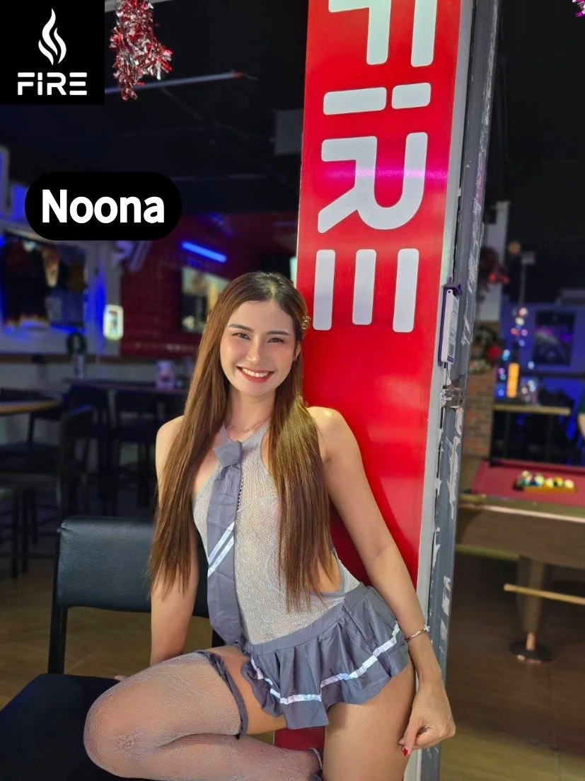 A young woman with long brown hair, smiling, and wearing a schoolgirl outfit with a gray tie, sitting on a chair in a bar or arcade. The background has neon lights, pool tables, and arcade games. There is a red vertical sign with white letters and a 