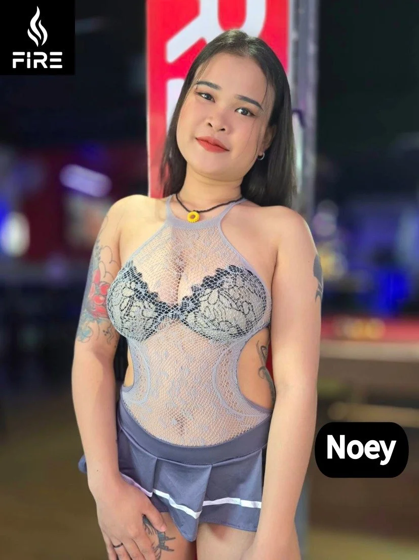 A woman with tattoos wearing a sheer lace top and shorts, standing in front of a blurred background at night with a Fire sign and red and white signs.