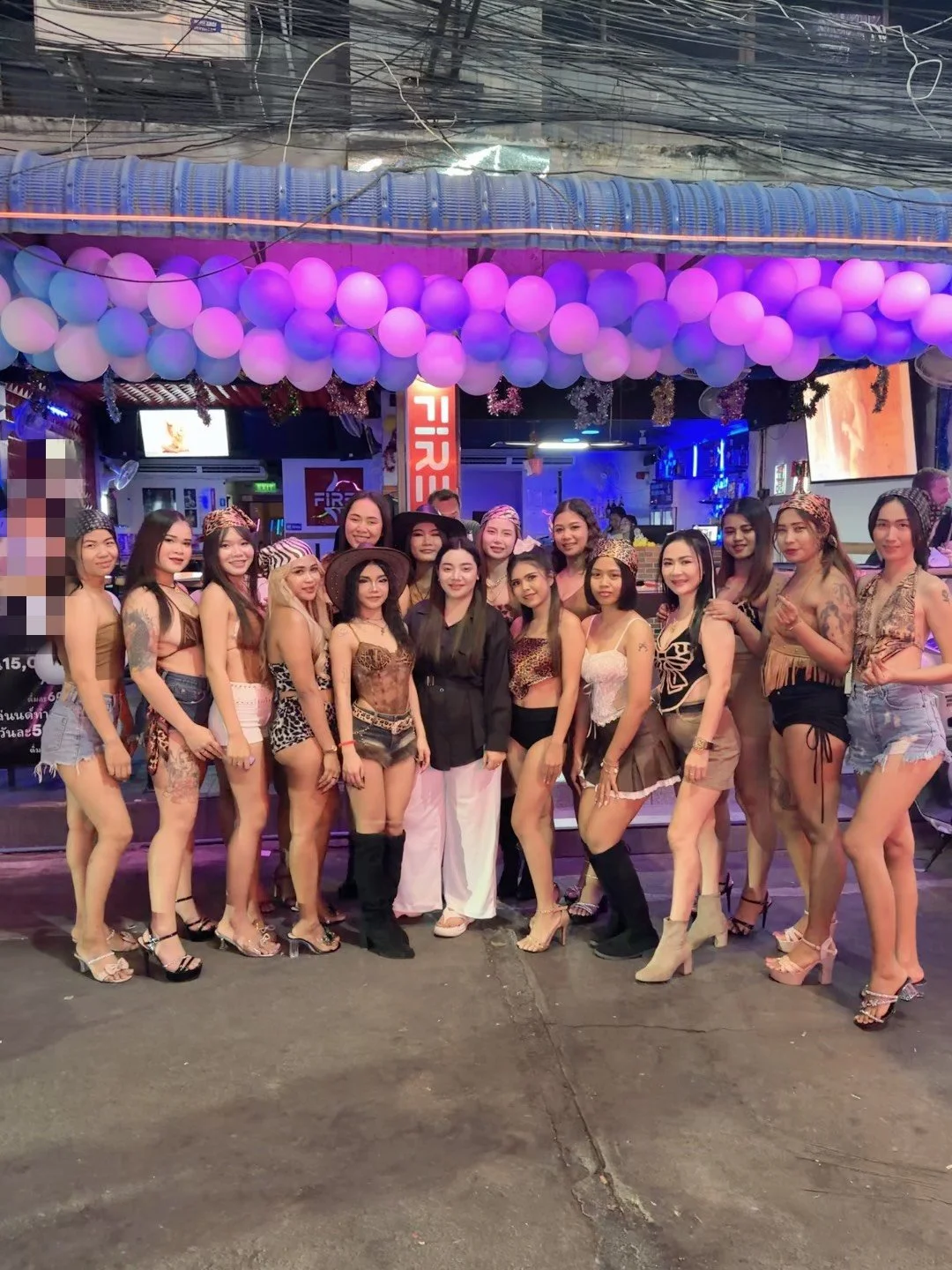 A group of women in various outfits posing in front of a decorated bar or club with purple balloons and neon lights.