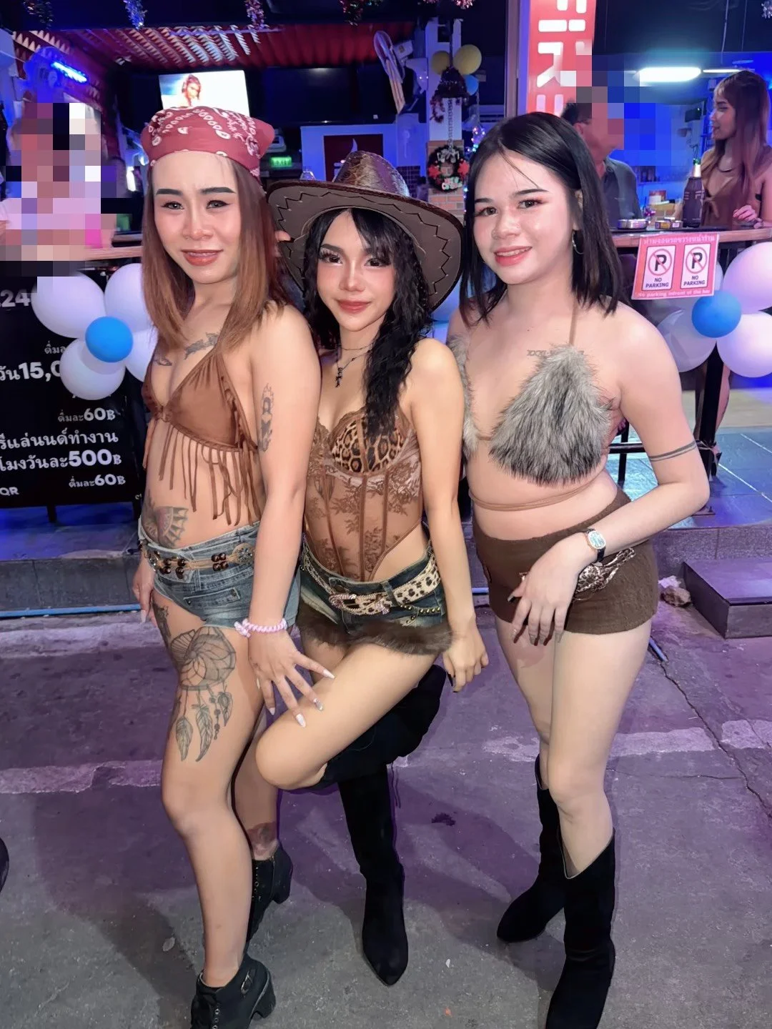 Three women in costumes posing together at a nightlife event, with a colorful decorated background and neon lights.