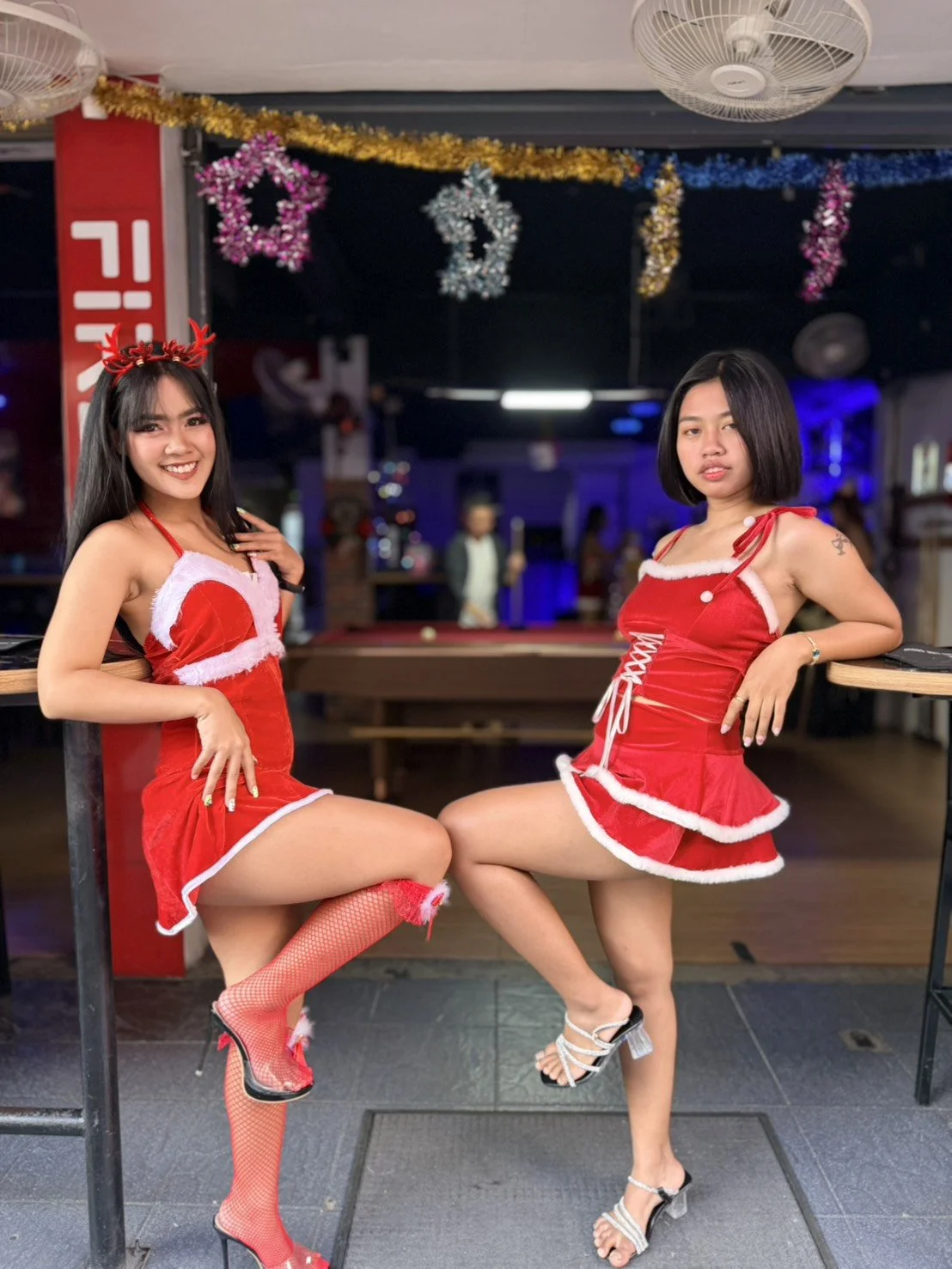 Two women in Christmas-themed costumes posing indoors with holiday decorations hanging from the ceiling.