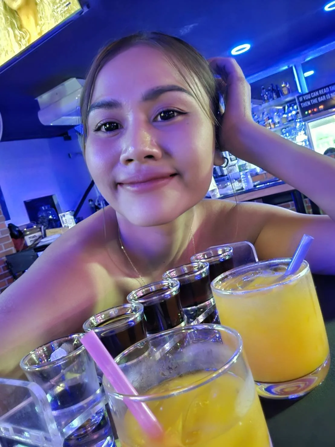 A young woman smiling at the camera in a bar, with several glasses of orange-colored drinks in front of her.
