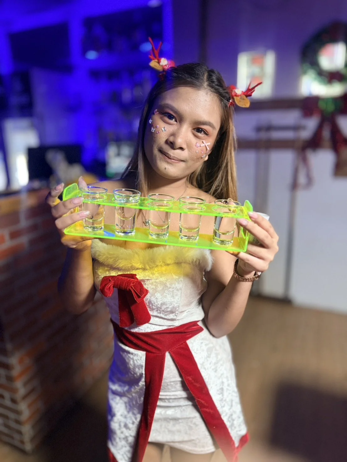 Woman dressed in holiday-themed outfit holding a tray of shot glasses in a festive setting.