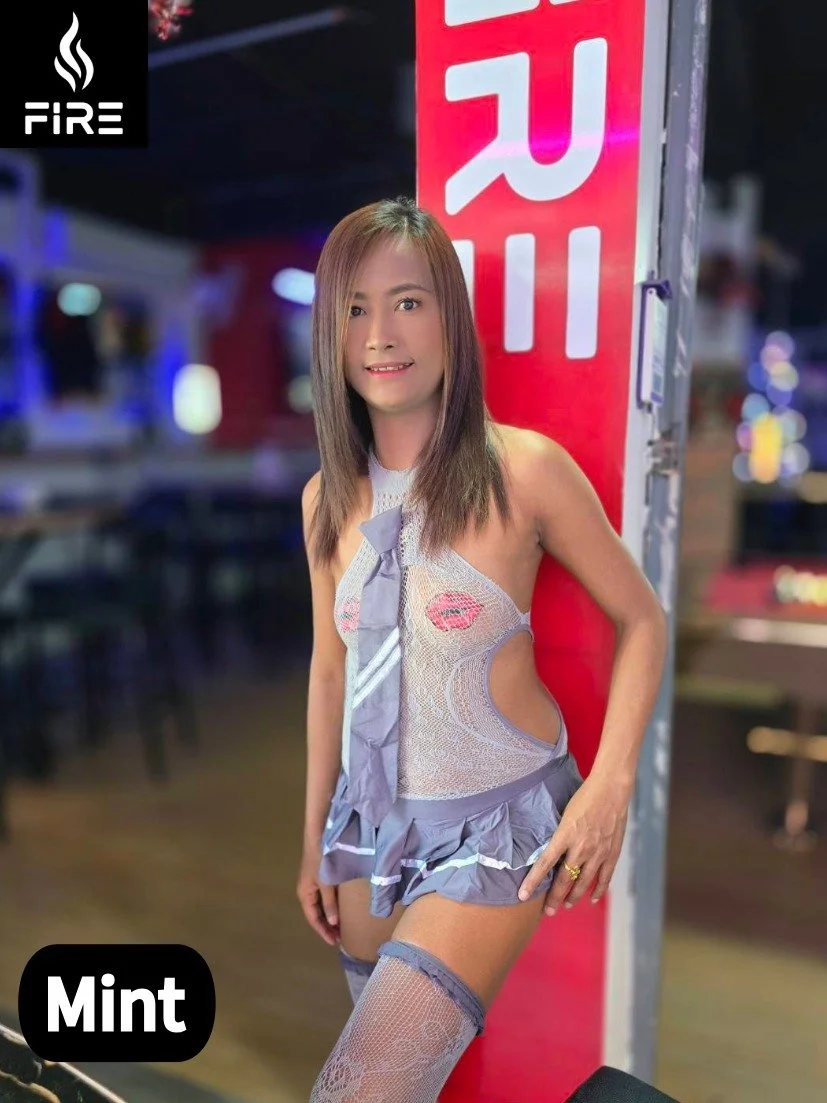 A woman with shoulder-length brown hair wearing a revealing fishnet lingerie costume with a small skirt, thigh-high stockings, and a tie, standing next to a red sign at an indoor event or club with blurred colorful lights in the background.