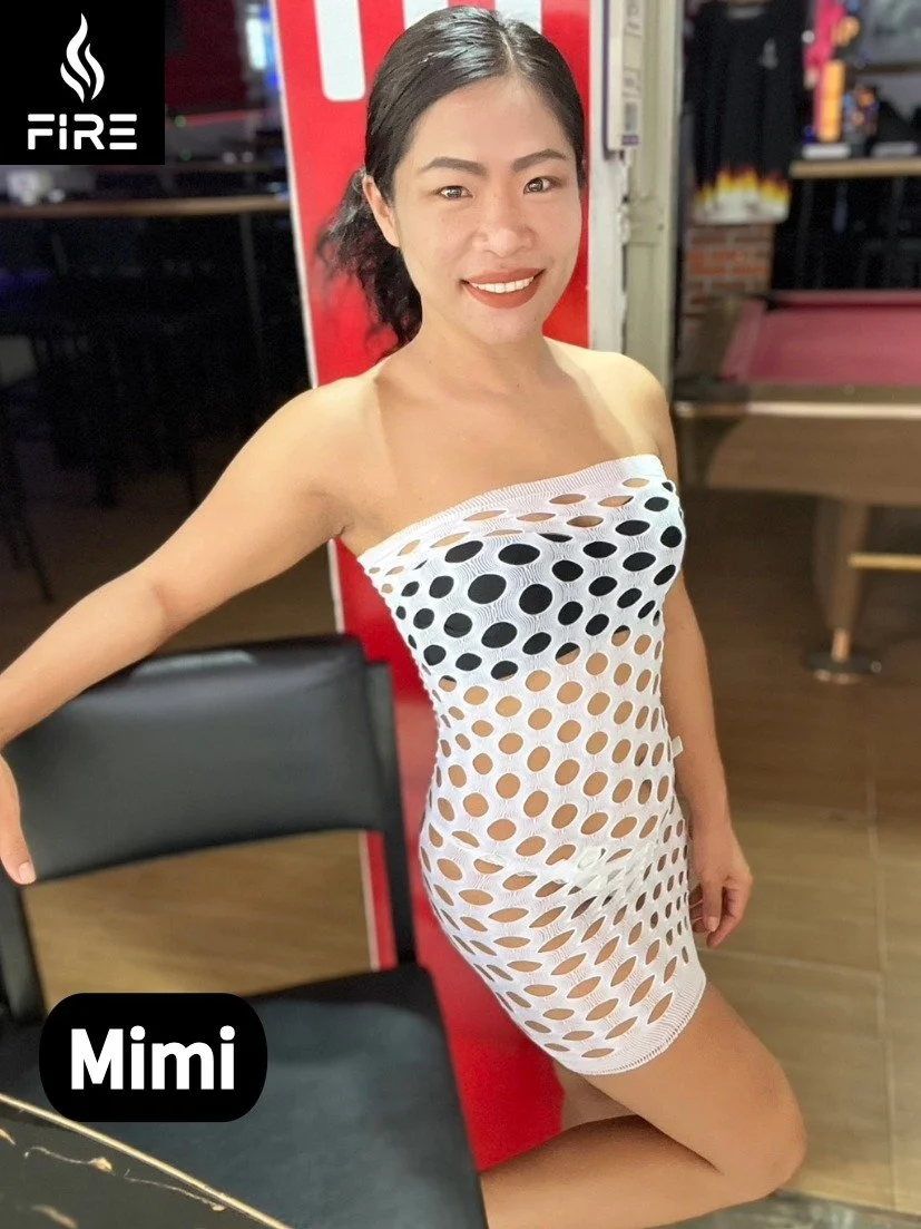 A woman with dark hair, smiling, wearing a strapless white dress with large black and beige dots and circular cutouts, posing indoors in a room with a pool table in the background.