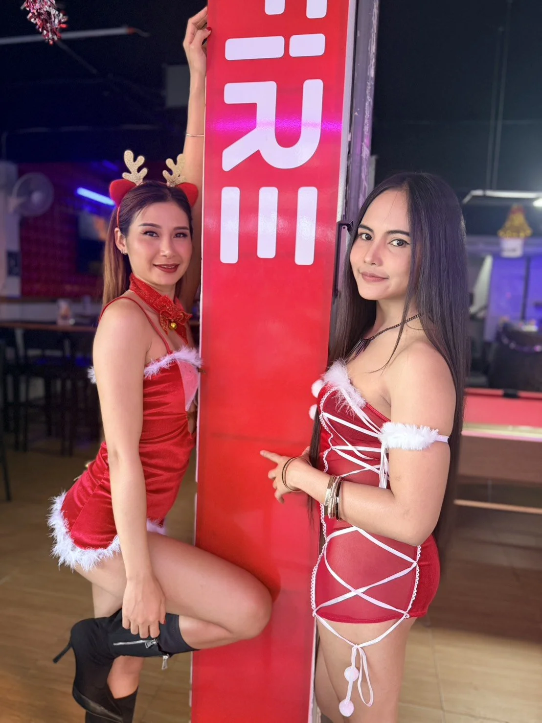 Two women dressed in Christmas-themed outfits posing next to a red vertical sign at a party or event.
