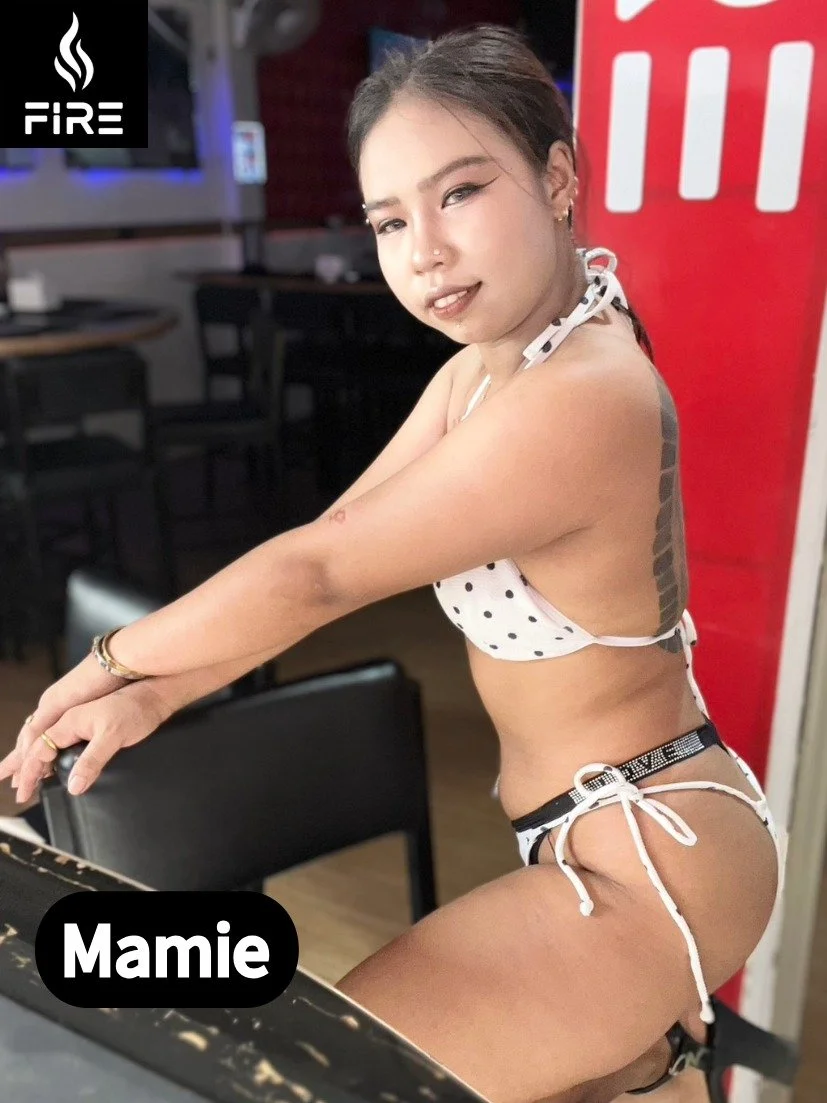 A woman with dark hair styled in a side braid, wearing a polka-dot bikini, sitting at a table in a restaurant or cafe. She has a tattoo on her back and is smiling slightly at the camera.