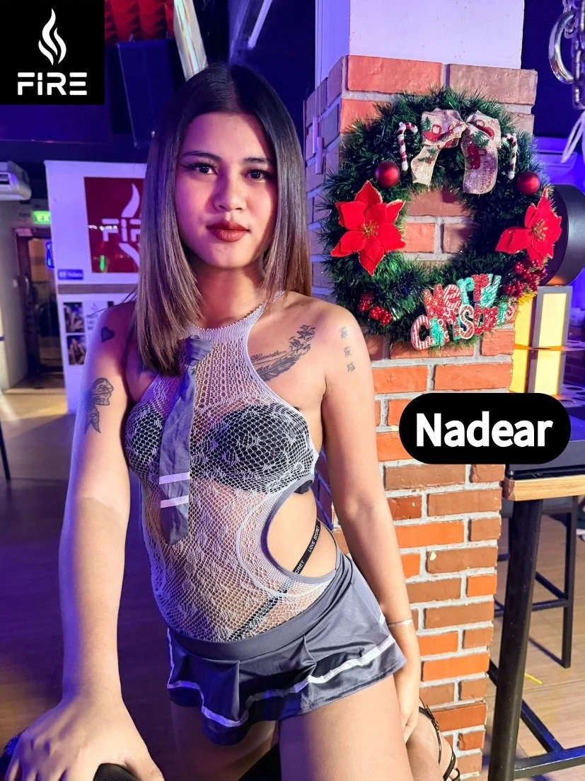 A woman with shoulder-length hair wearing a see-through mesh top and a short skirt, standing indoors near a Christmas wreath on a brick wall.