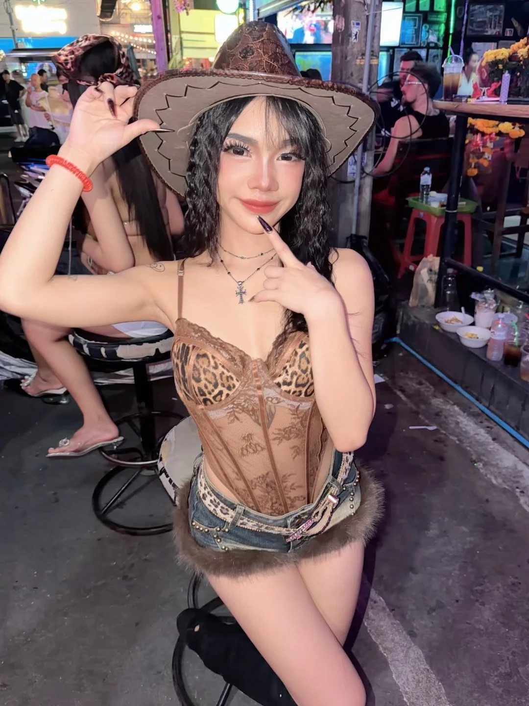 A young woman with long black hair, wearing a cowboy hat and a leopard-print corset top, poses with a finger on her cheek at an outdoor event or street fair at night. She has layered necklaces and is standing in front of a busy, colorful background w