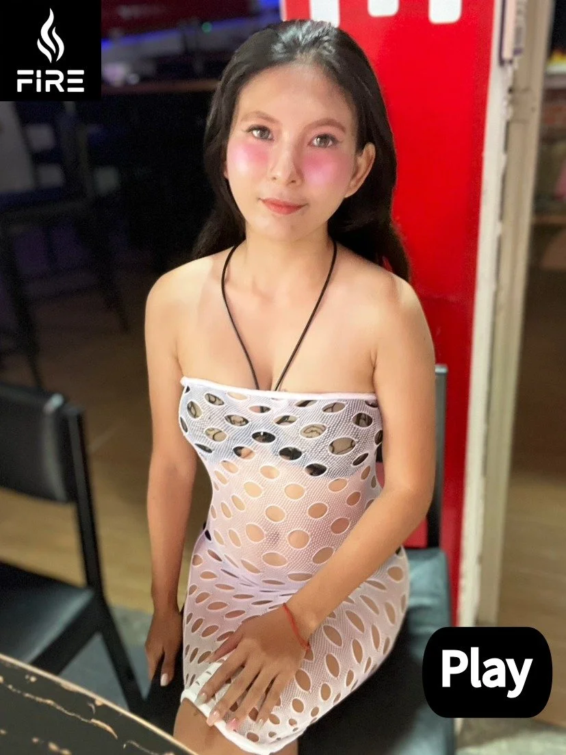 A young woman with long dark hair and pink blush on her cheeks, wearing a white see-through dress with large circular cutouts, sitting on a black chair in a dimly lit indoor setting.