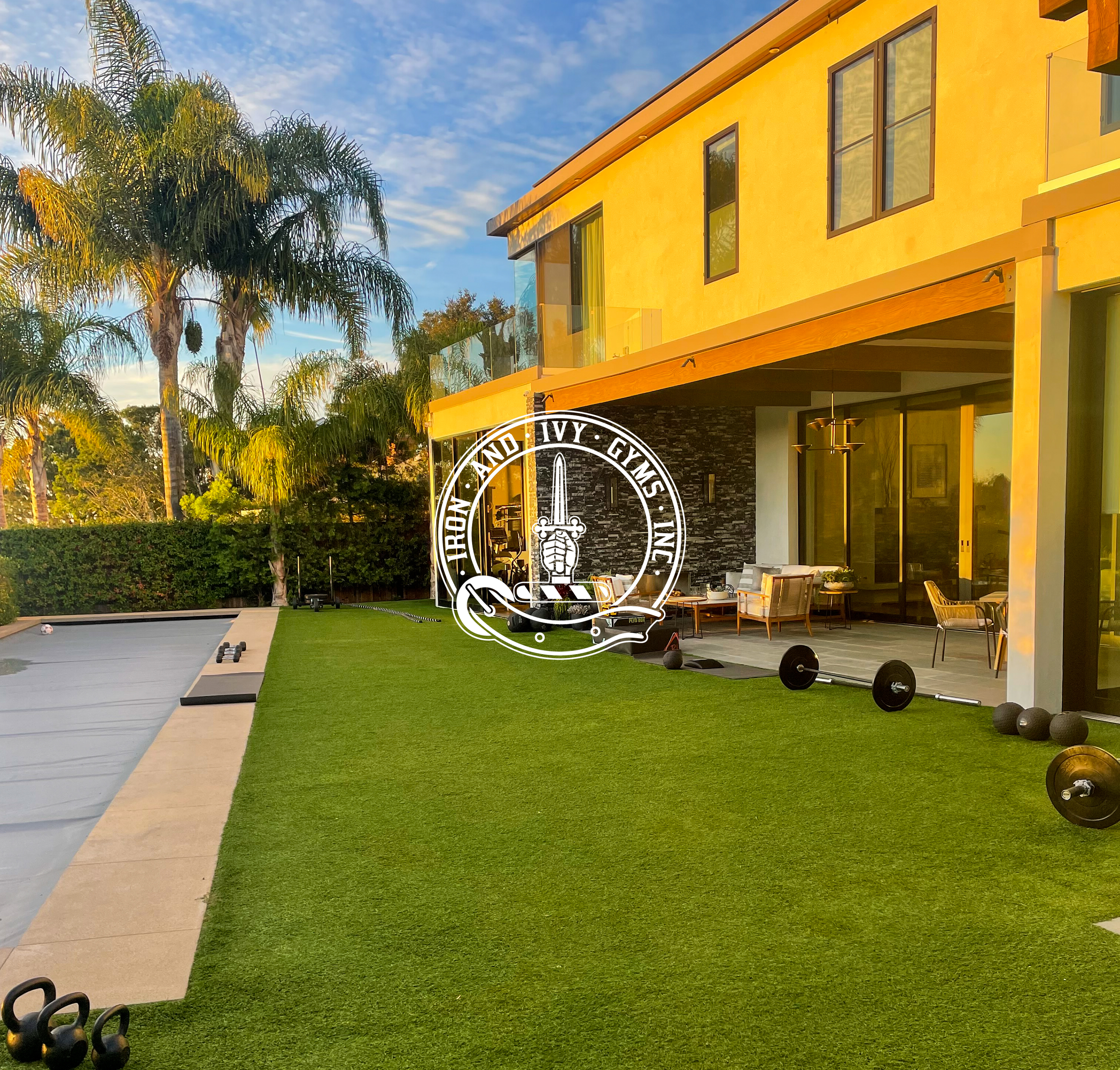 Modern yellow house with outdoor gym equipment on well-maintained artificial grass, alongside a patio with seating area, surrounded by tall palm trees and lush greenery under a blue sky.