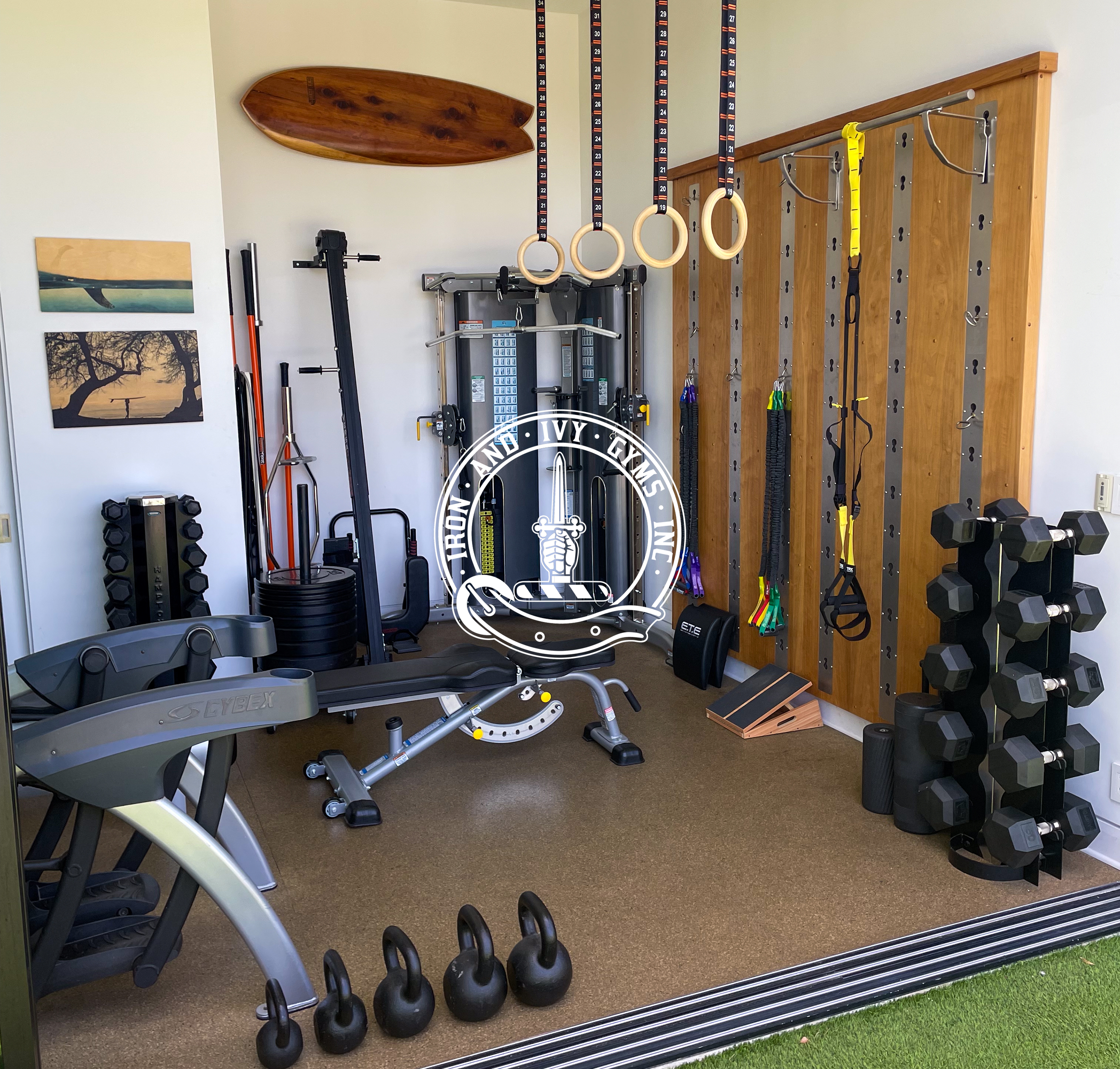 Home gym with weight rack, kettlebells, training equipment, wooden wall with gymnastic rings and resistance bands, surfboard and artwork on the walls.