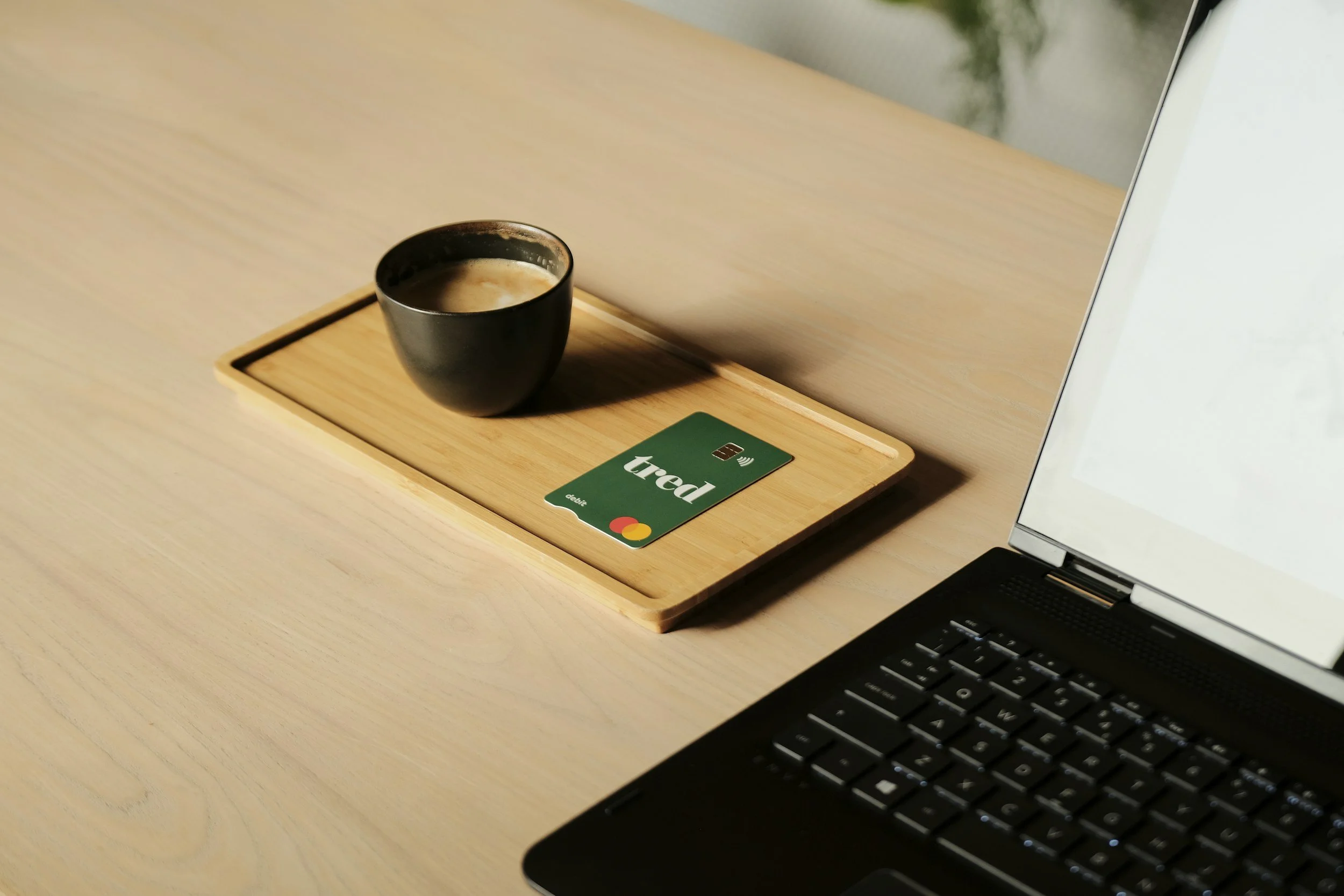 A black cup filled with coffee or latte on a wooden tray, with a green credit card next to it, placed on a light wooden desk next to a laptop.