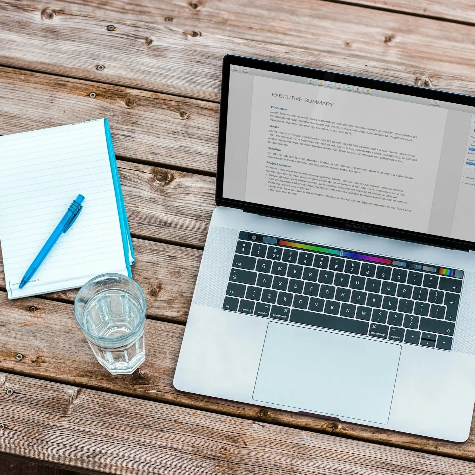 A laptop with a document titled 'Executive Summary' on the screen, a notebook with a blue pen, a glass of water, all on a wooden table.